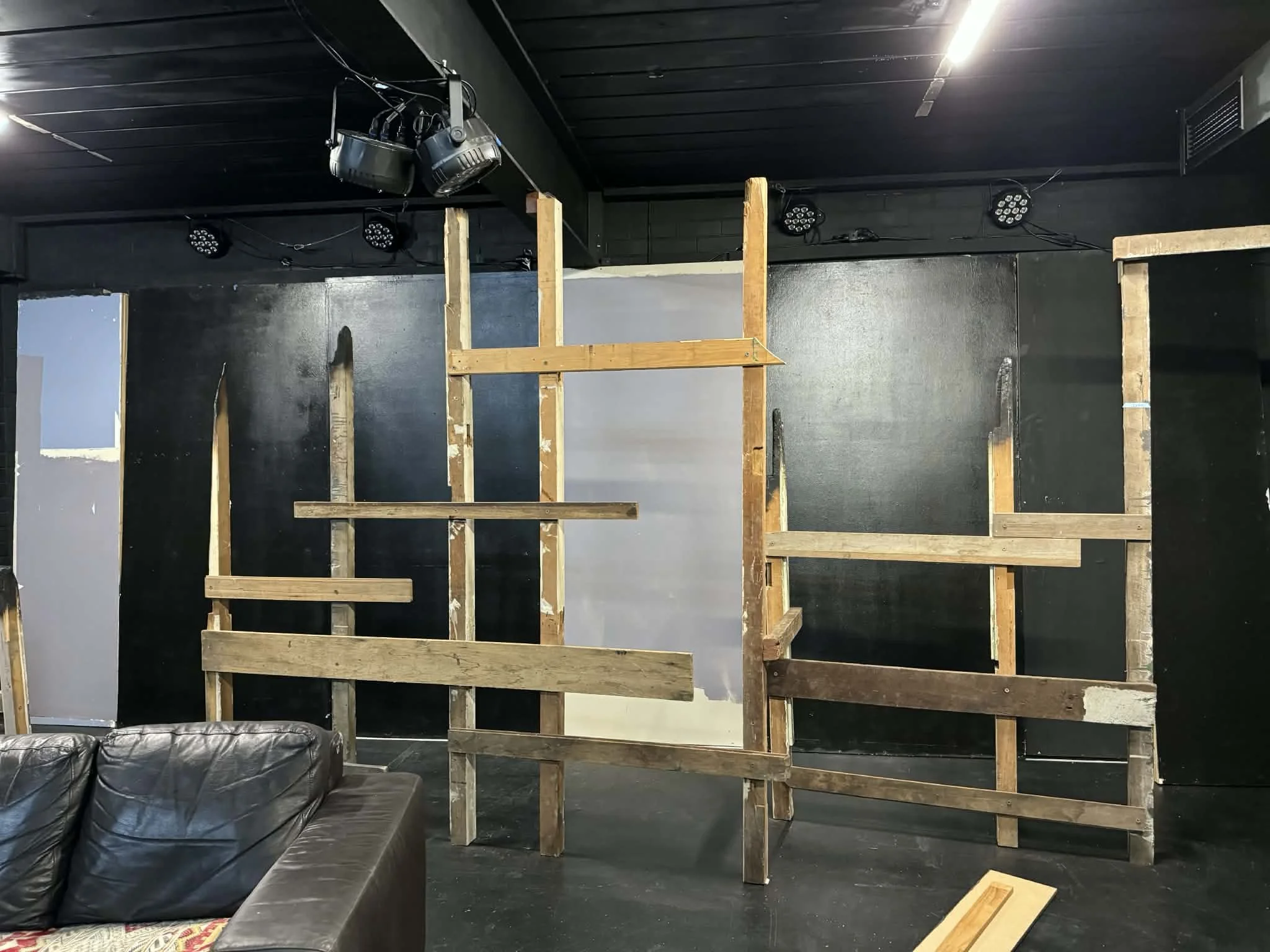 Interior of a stage setup with wooden frames for a backdrop or scenery, black wall and ceiling, stage lights and speakers, and a black leather sofa in the foreground.