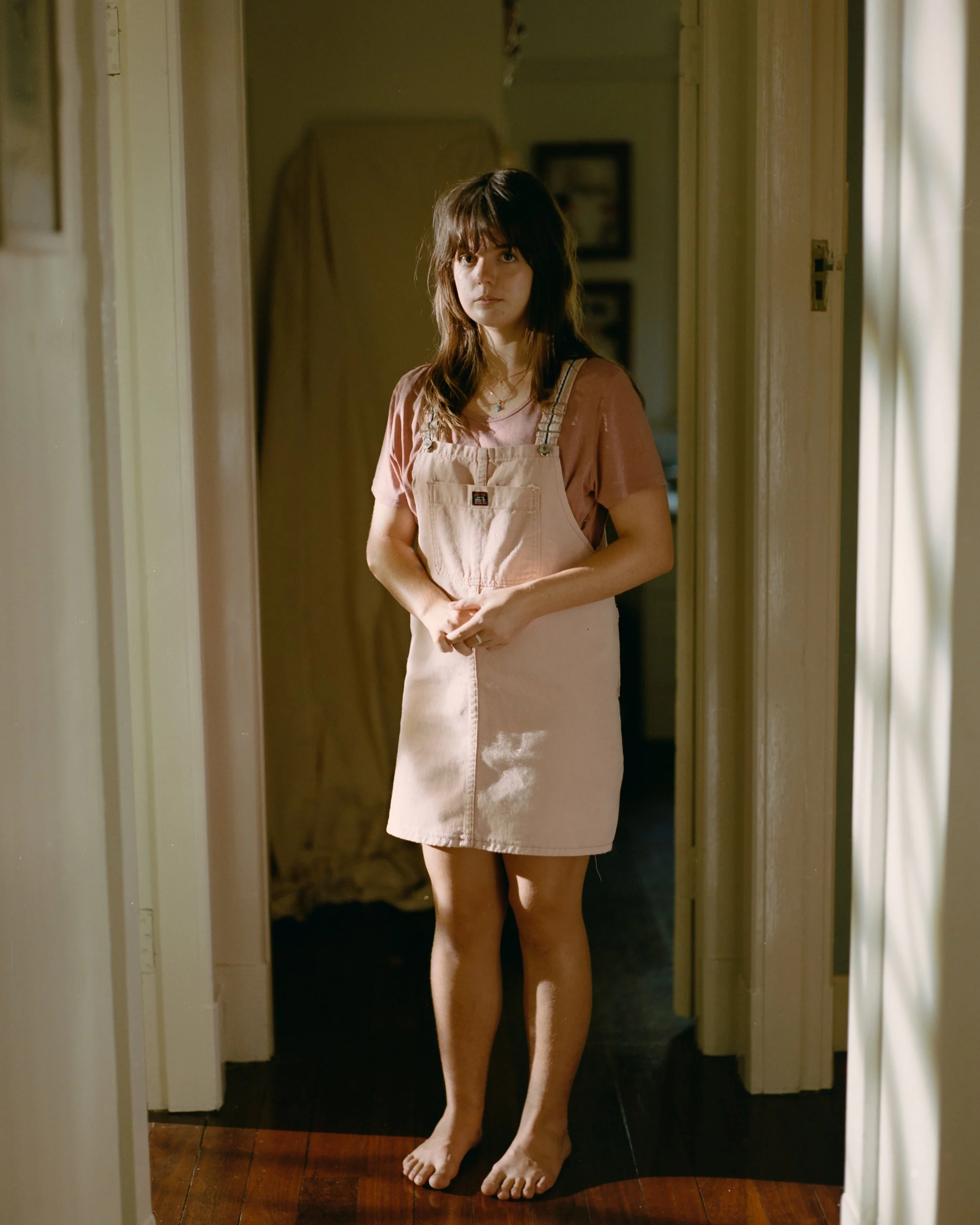 A young woman with brown hair and bangs, wearing a pink t-shirt and a beige overall skirt, standing barefoot in a hallway with natural light.