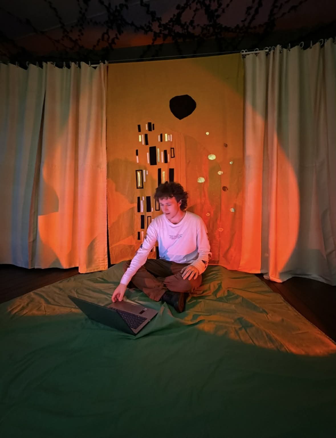 Person sitting cross-legged on a green mat in front of a laptop, with curtains and an abstract orange and black backdrop behind him.