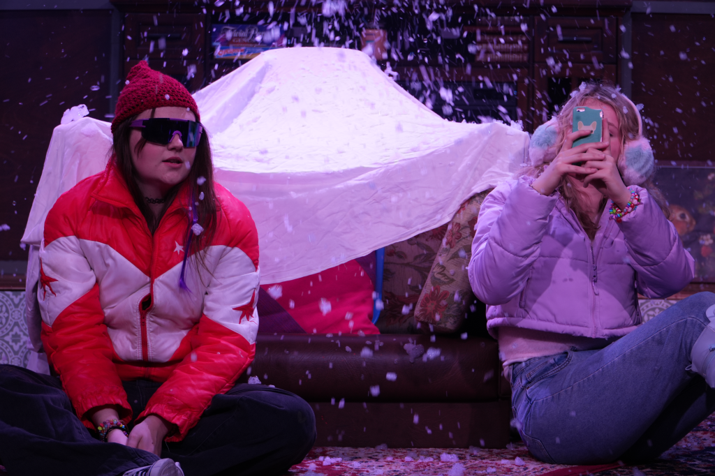 Two girls sitting on a couch, one wearing sunglasses and a red hat, the other taking a photo, with a white sheet and snow-like effect in the background.