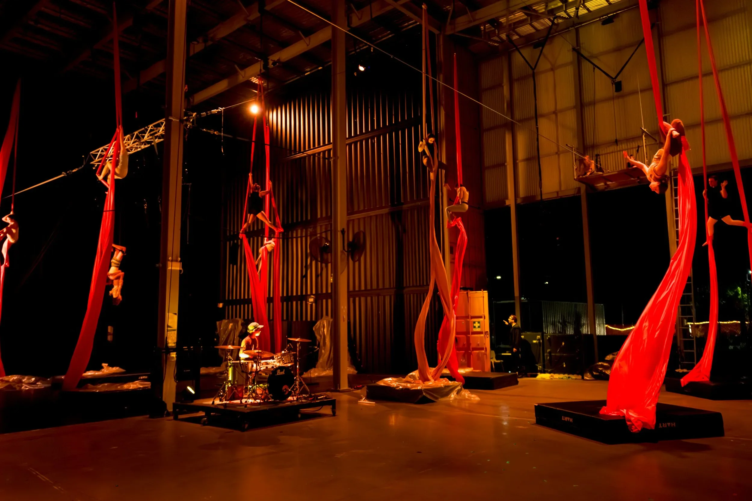 Aerial silk performers hanging from red silk fabrics in a dark indoor performance space, with musicians playing drums on a stage in the foreground.