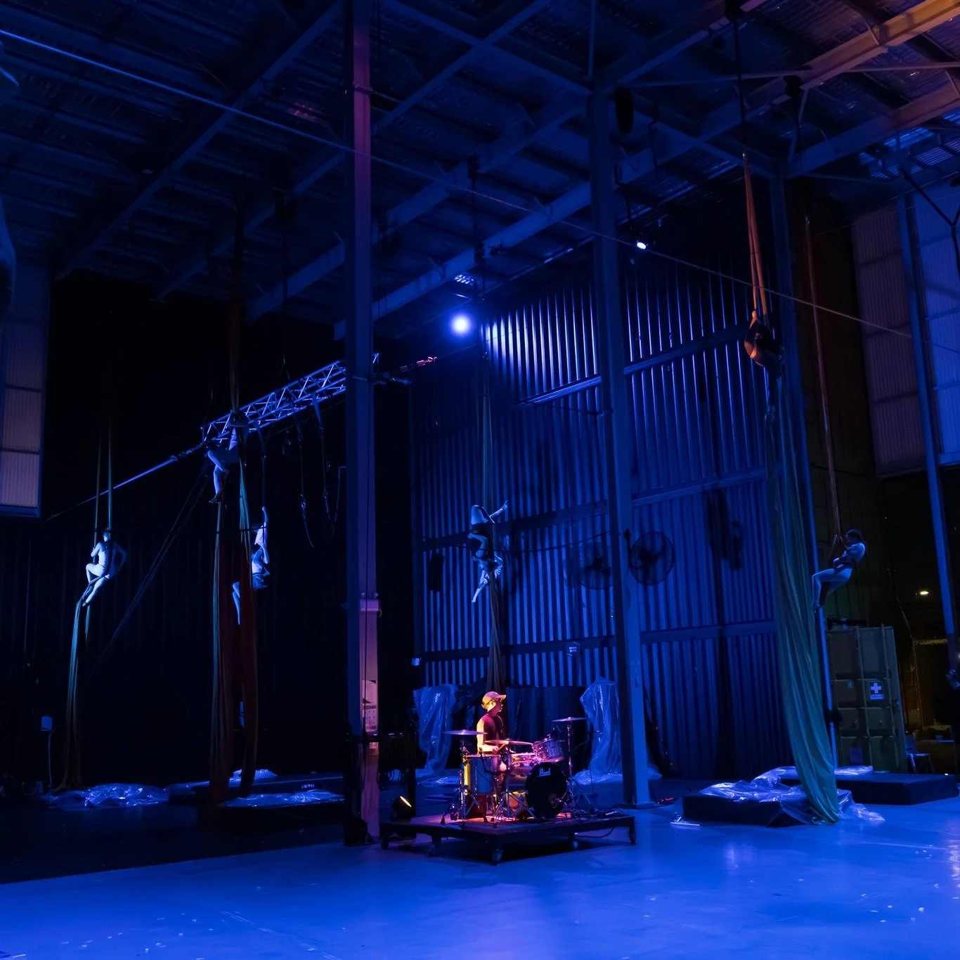 Aerial performers practicing in a dimly lit theater with blue lighting, suspended in the air using silks and ropes, and a musician playing drums on stage.