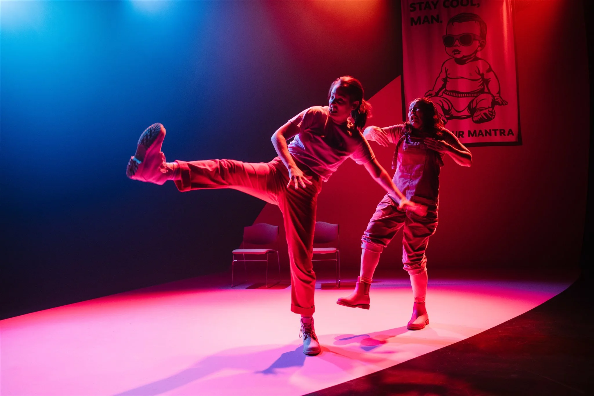 Two women dancing on a stage with red and purple lighting, one lifting her leg and the other touching her collarbone, with chairs and a poster in the background.
