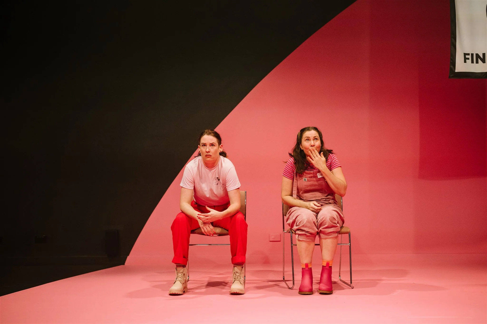 Two women sitting on chairs on a stage with a pink and black background, one covering her stomach and the other with her hand over her mouth, both looking surprised or shocked.