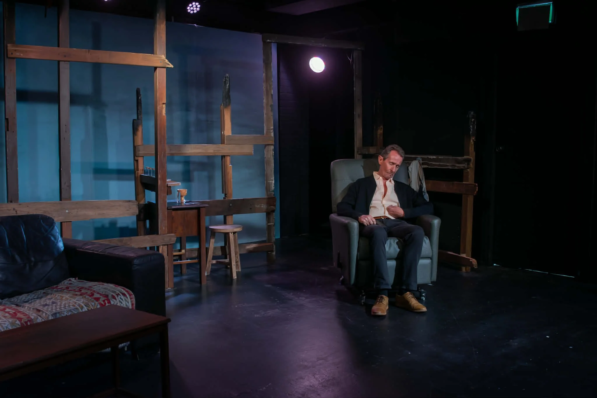 A man in business attire seated in an armchair on a dark stage, with a wooden framework and furniture around him, looking at his phone.