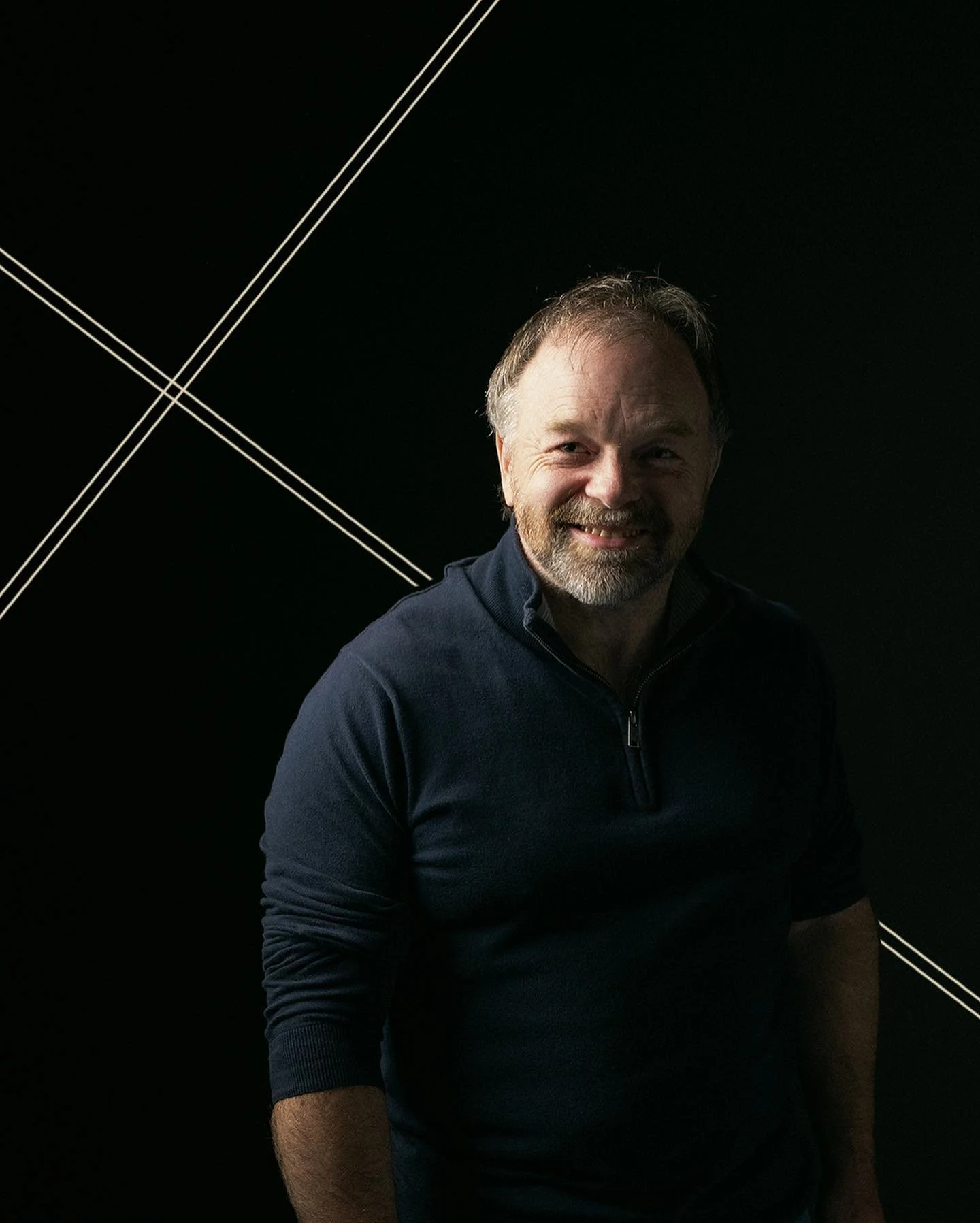A smiling middle-aged man with a beard and mustache, wearing a dark zip-up sweater, stands in front of a black background with white diagonal lines.