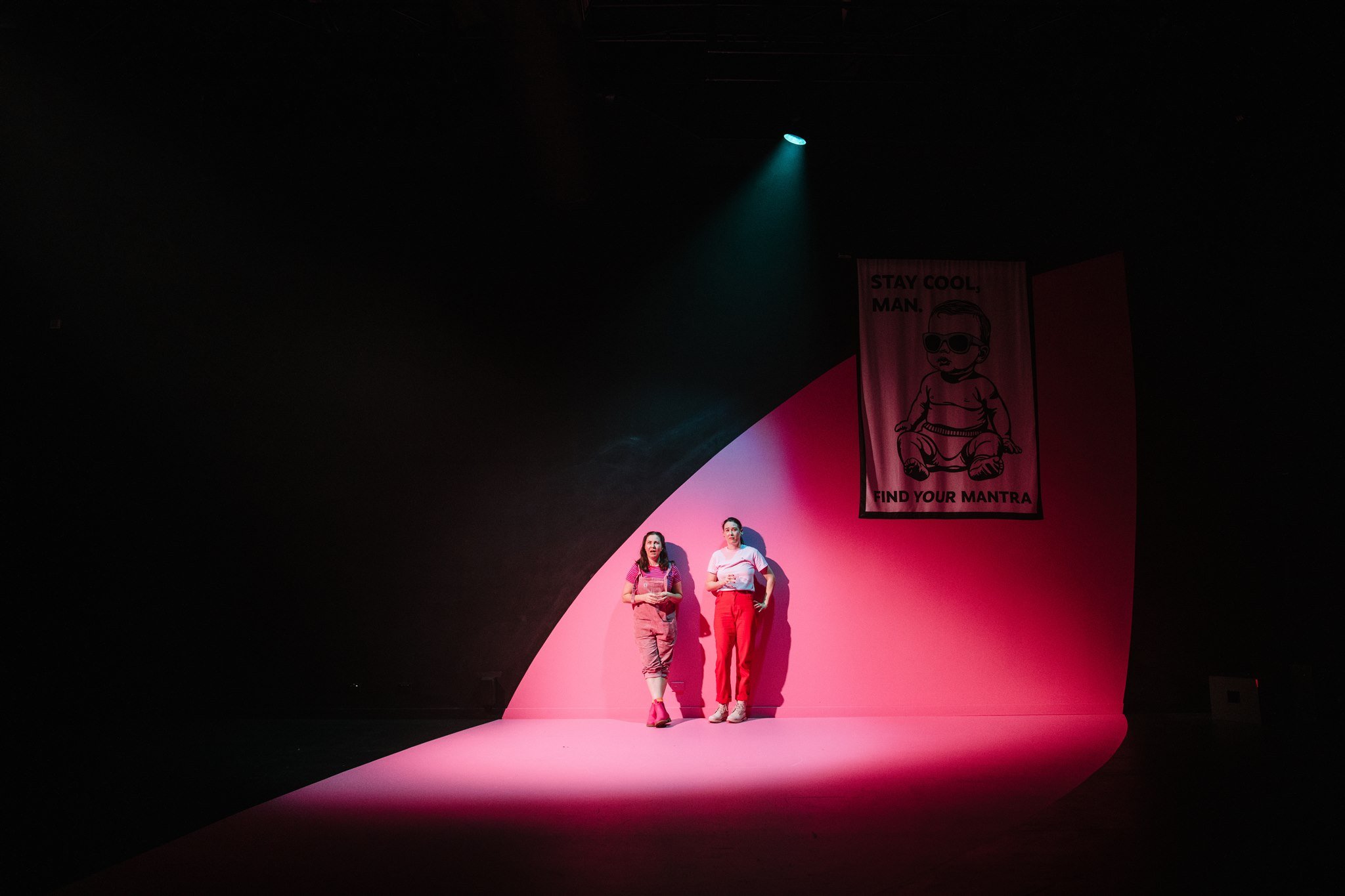 Two people standing on a pink stage with a black background, illuminated by a spotlight. A banner with a cartoon character and the text "Stay Cool Man. Find Your Mantra" hangs above them.