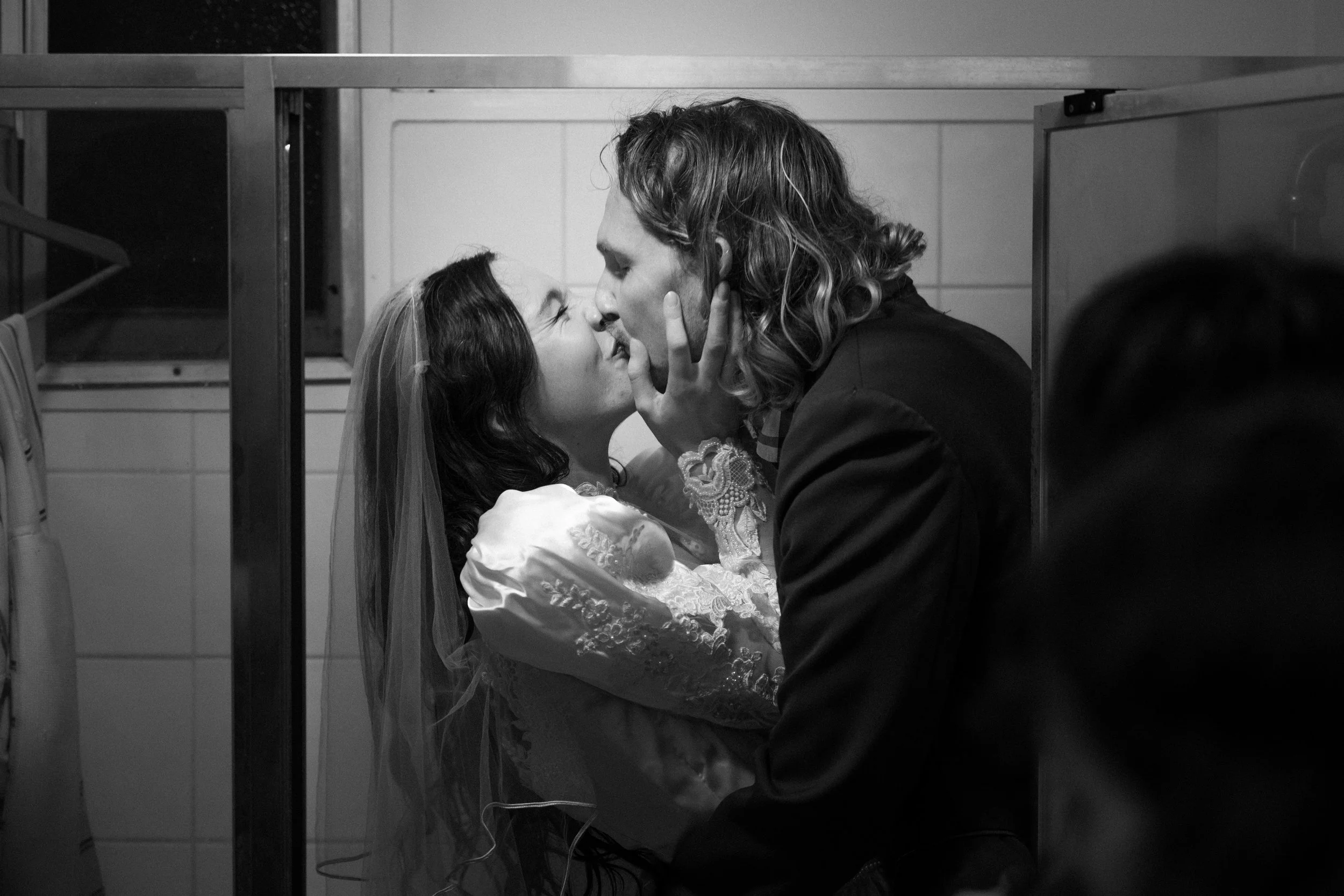 A couple sharing a kiss, possibly on their wedding day, in a black and white photo in what appears to be a confined space like a bathroom or closet.