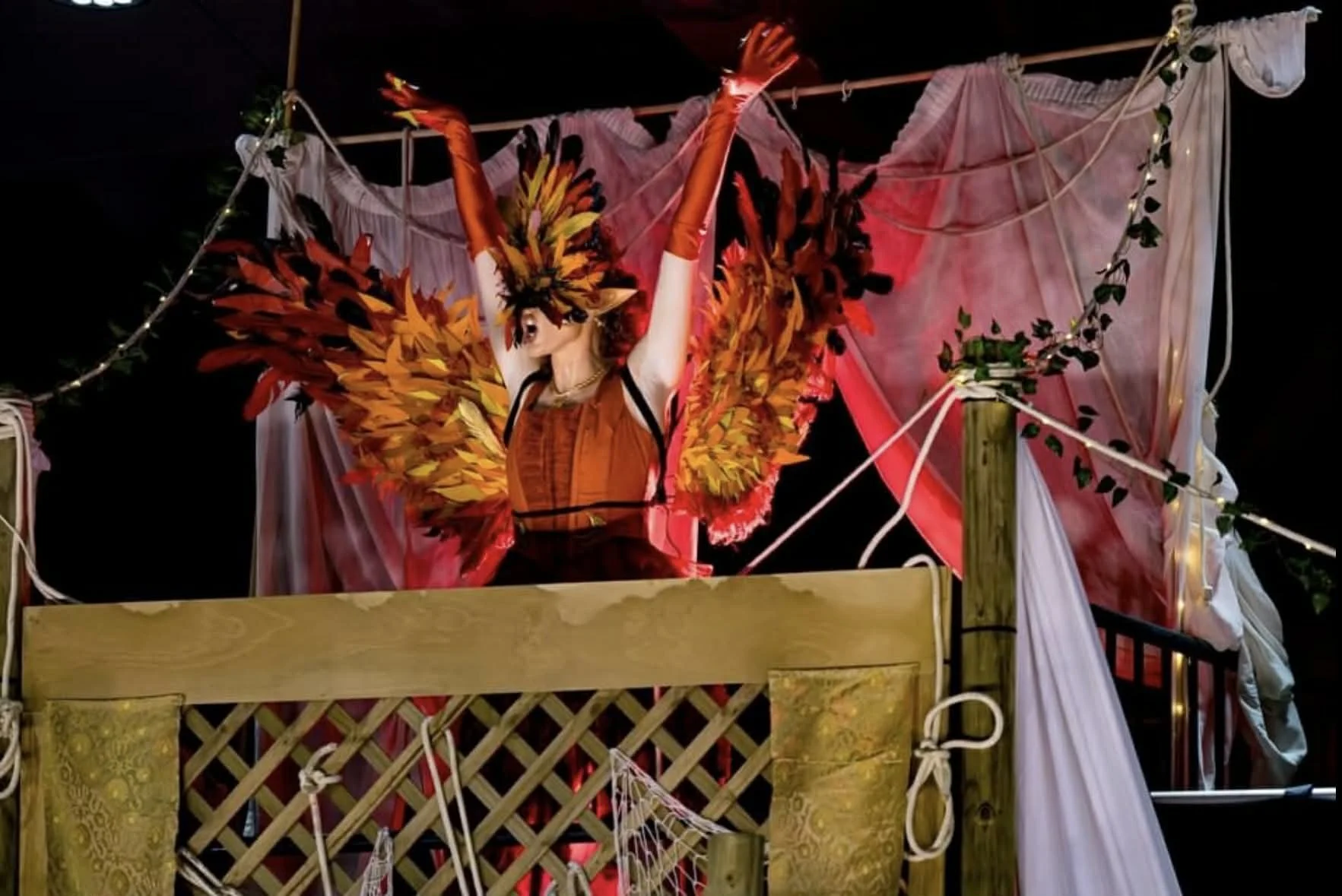 A figure dressed as a performer or fairy with large, colorful yellow, orange, and red feathered wings, raising arms and standing on a decorated float or stage with pink and white fabric and string lights at night.