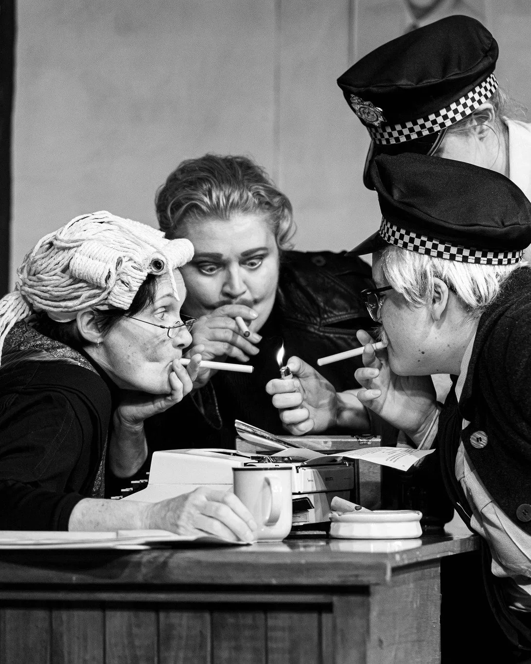 Four women, two with police hats, one with hair rollers, all smoking cigarettes and sharing a lighter around a table in black-and-white photo.