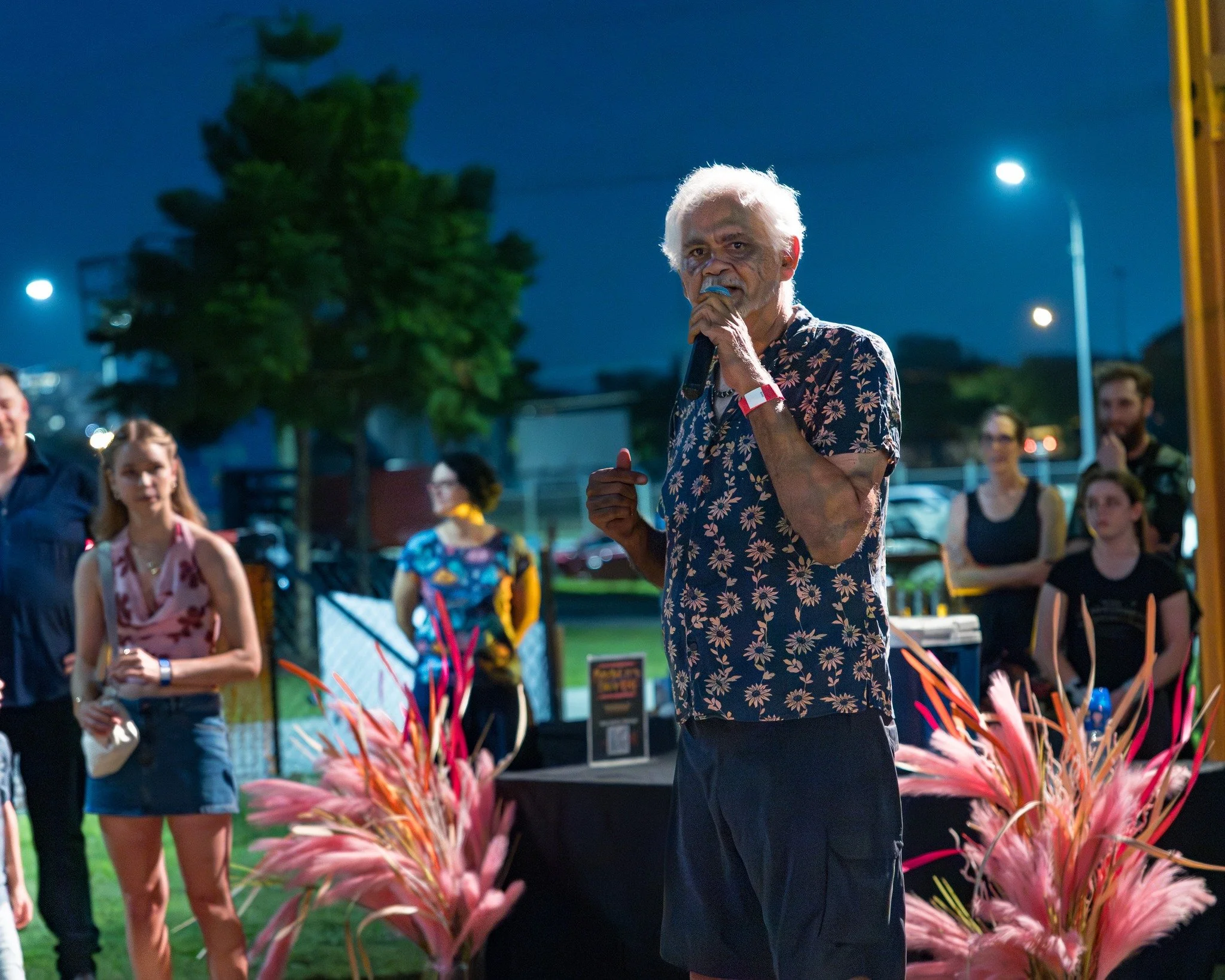 An elderly man with white hair speaking into a microphone at an outdoor event during nighttime, surrounded by people and pink decorative plants.