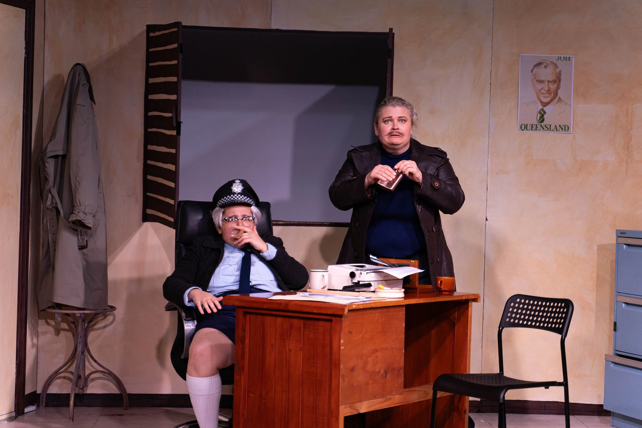 A woman and a man in office, woman standing with surprised expression, holding a small item, man seated at desk with glasses, police hat, and snack, underwear visible, office cluttered with papers and boxes, Australian-style tan wall, poster of a man