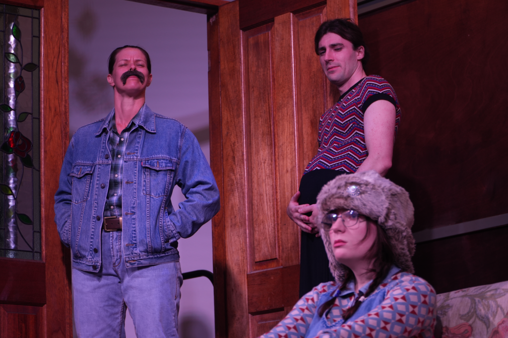 Three individuals in a room with wooden walls: a man with a mustache in a denim jacket, another man with dark hair in a patterned shirt and holding his stomach, and a woman with glasses and a furry hat sitting on a couch.