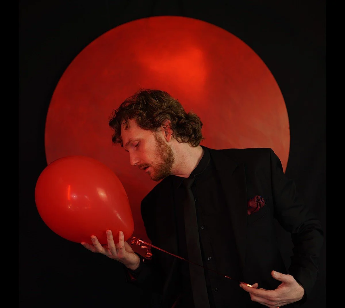 A man with wavy hair and a beard in a black suit holding a red balloon against a backdrop featuring a large illuminated red circle.