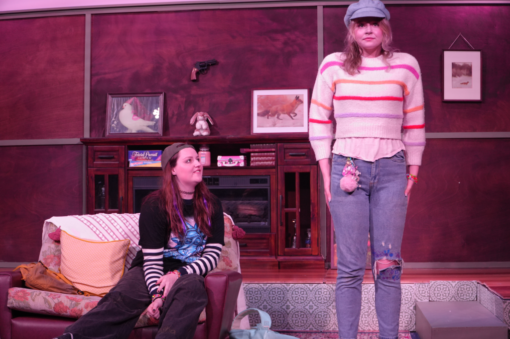 Two women in a living room with wood-paneled walls, decorated with framed pictures and a gun mounted on the wall. One woman is sitting on a floral-patterned couch wearing a black and striped shirt, while the other stands in front of her wearing a pas