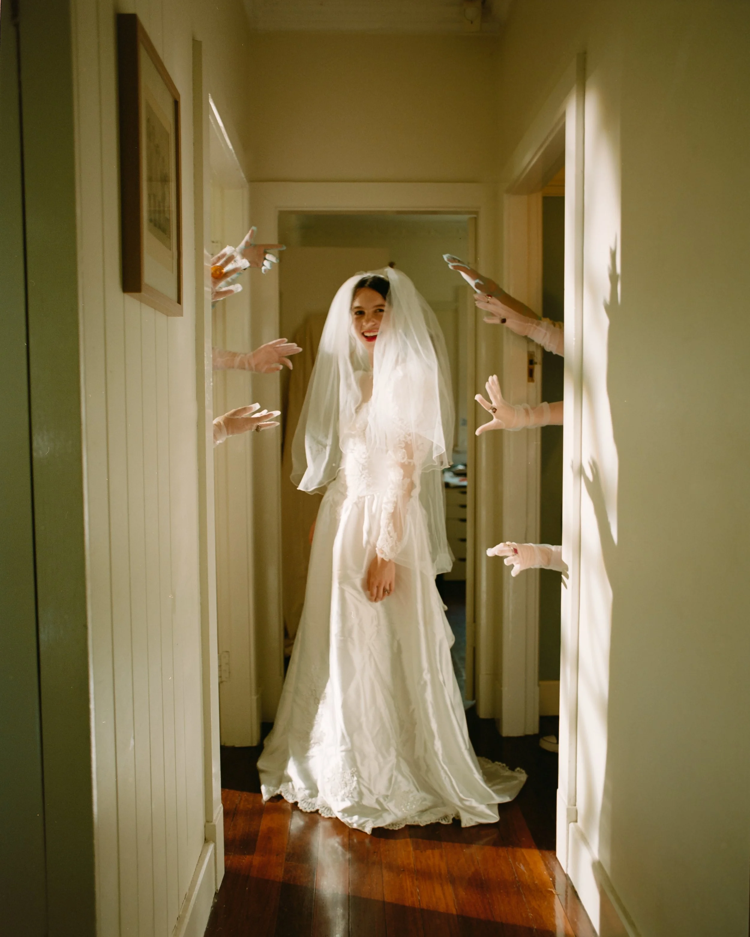 Bride in a white wedding dress and veil smiling as she stands in a hallway, surrounded by ghostly hands reaching out toward her.