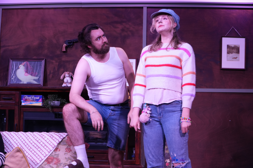 A man with a beard and long hair wearing a white tank top and shorts, sitting on furniture, looking at a woman. The woman has blonde hair, is wearing a colorful striped sweater, a cap, and jewelry, standing with a confident expression.