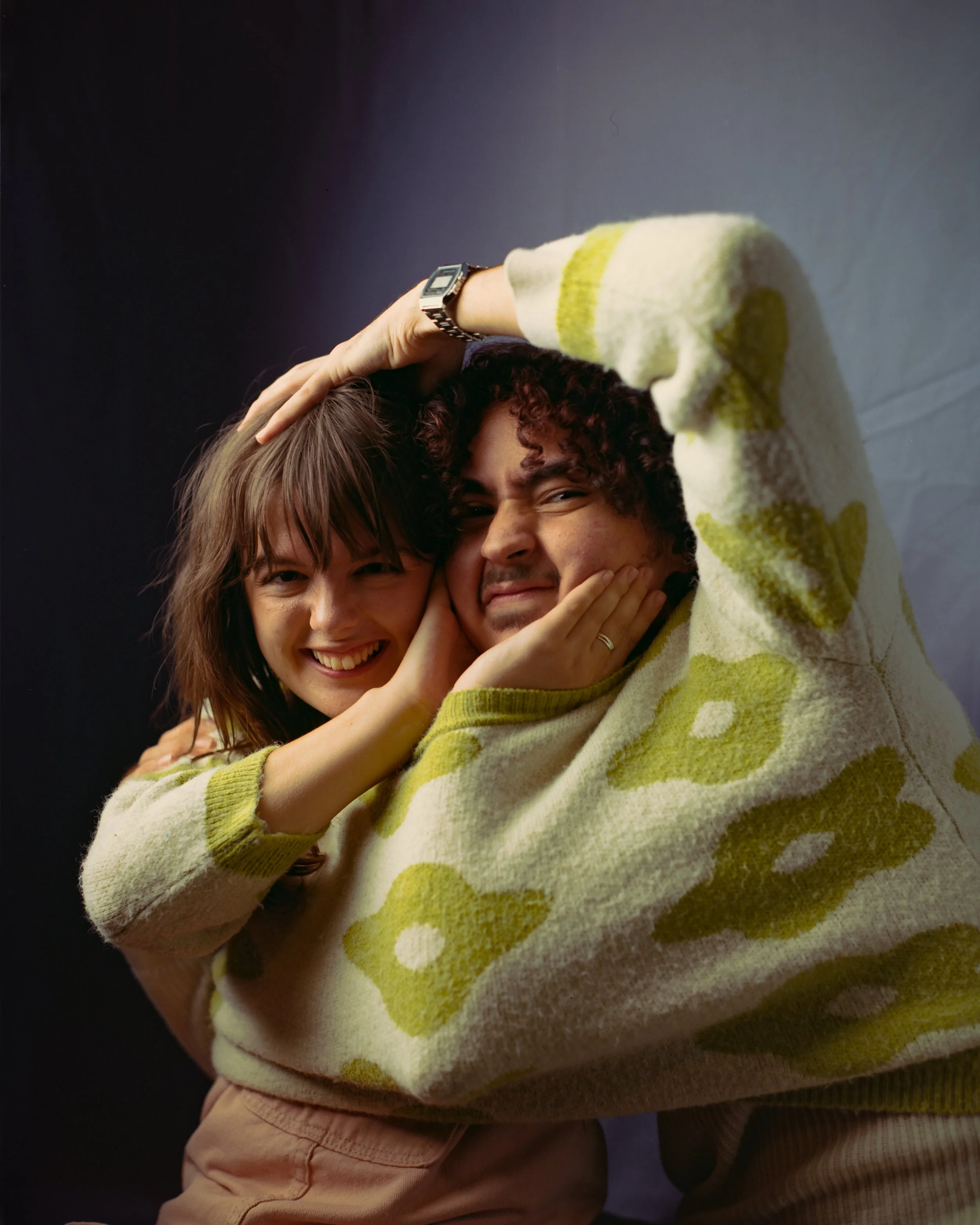 Two people, a woman with brown hair and a man with curly dark hair, hugging and smiling at the camera. The woman is holding the man's face with her hand, and the man is smiling with his eyes squinting. They are both touching each other's faces, and t