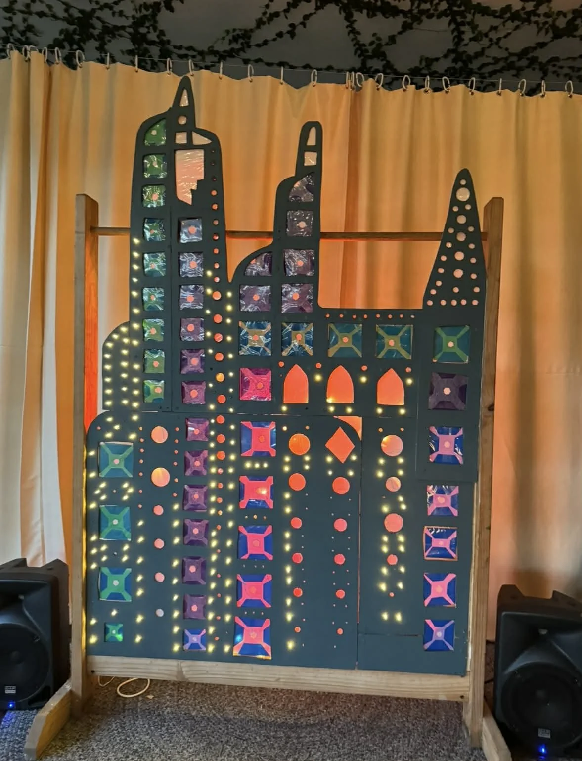 Decorative illuminated cardboard cutout of the Brooklyn Bridge at night, with colorful lighting and a dark background, flanked by beige curtains.