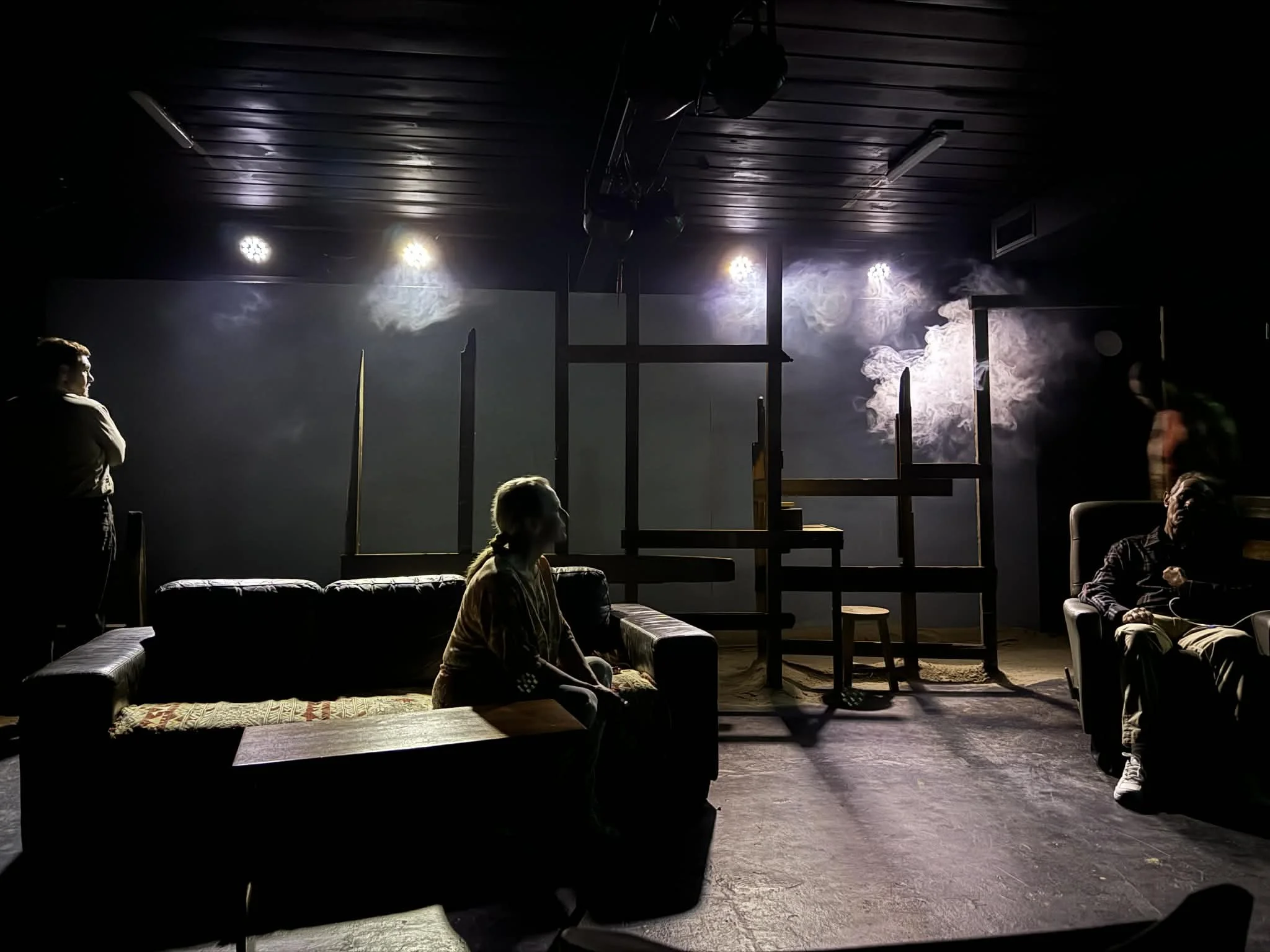 People sitting on sofas in a dimly lit room with stage lights and smoke, possibly a theater or performance space.