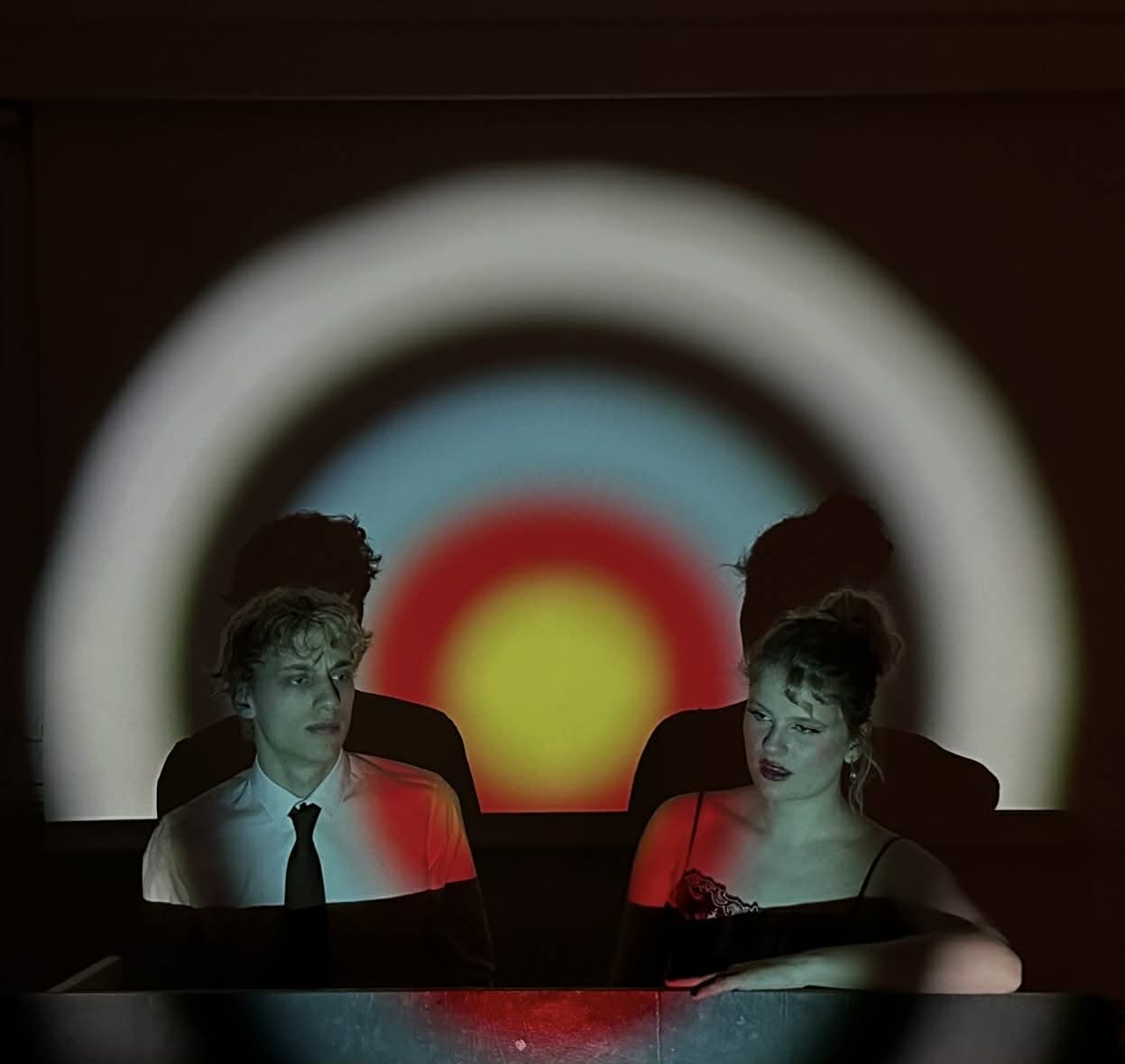 A man and a woman sitting at a table with a colorful concentric circle projection behind them, creating a dramatic and artistic scene.