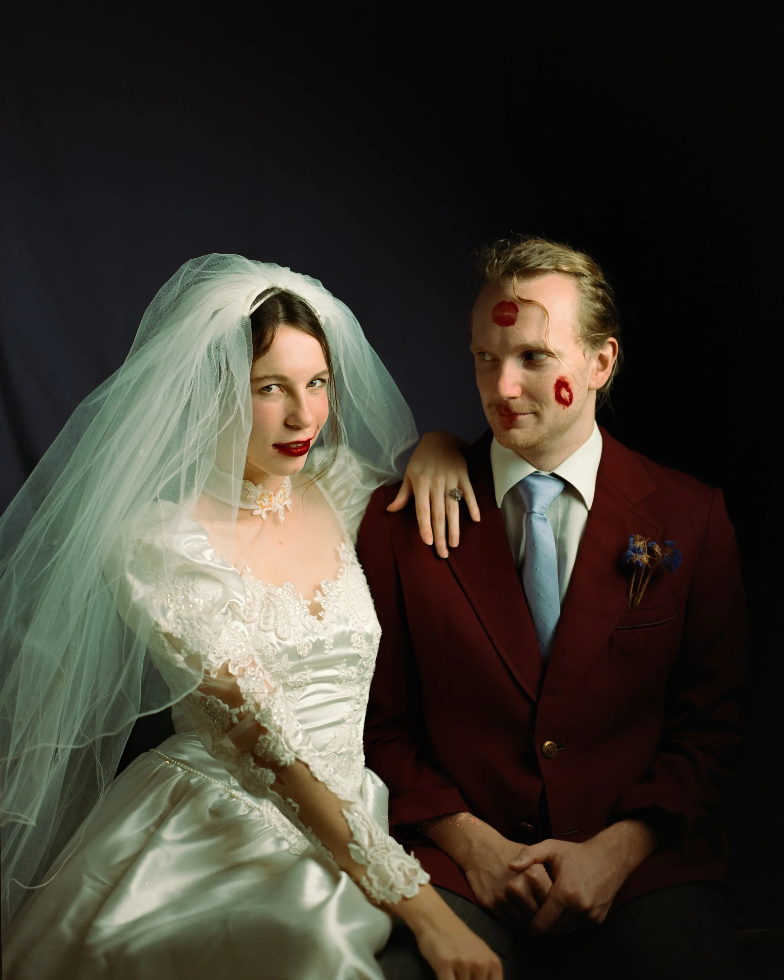 A woman in a wedding dress and veil with dark lipstick sitting next to a man in a burgundy suit with lipstick kiss marks on his face, against a dark background.