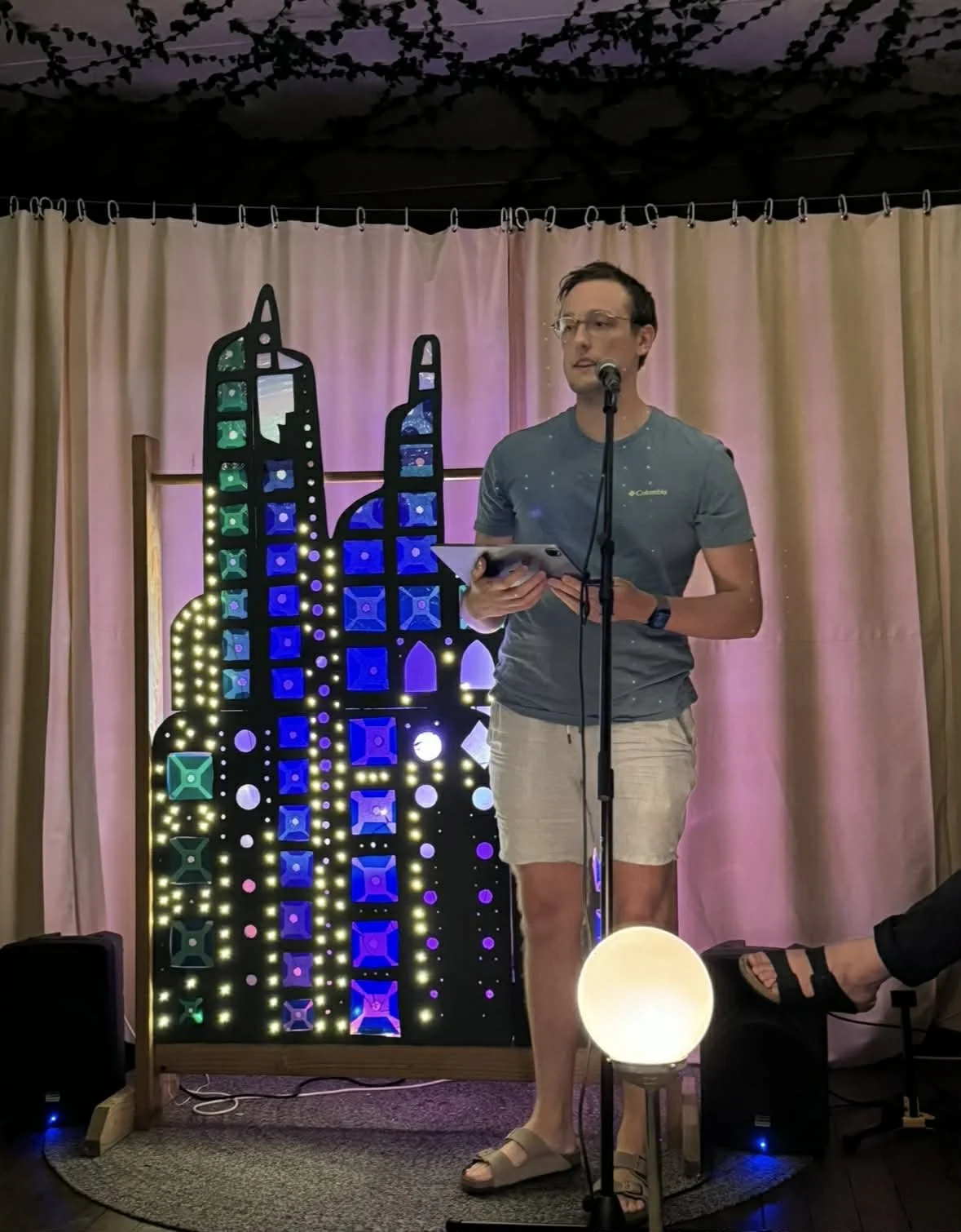 A young man standing at a microphone on stage, holding a tablet, with a cityscape-themed light display and pink curtains in the background.
