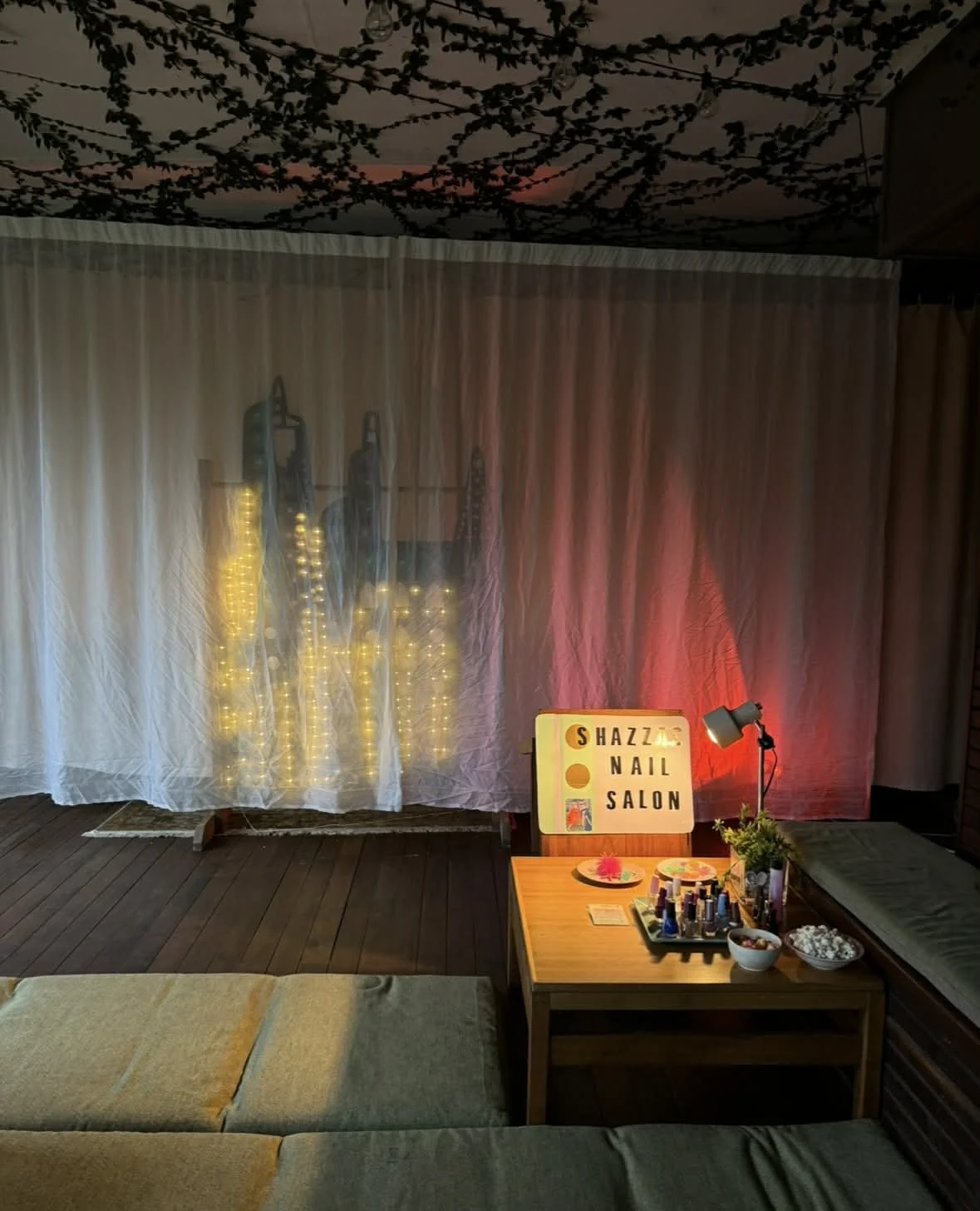 Decorative setup at Shazza Nail Salon with a sign, nail polish bottles, bowls of snacks, a small plant, a lamp, and a backdrop featuring fairy lights and curtains.