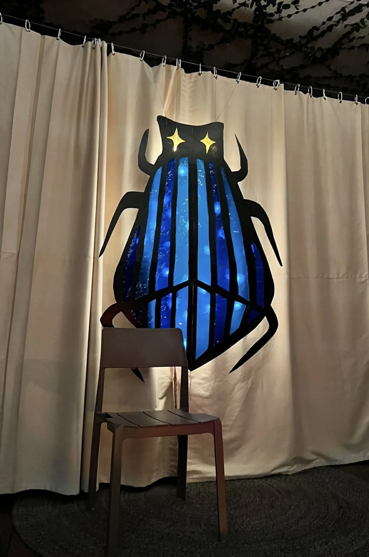 Large decorative beetle projected on a beige curtain, with black outlines, blue patterned interior, and yellow star-shaped eyes, with a wooden chair in front.