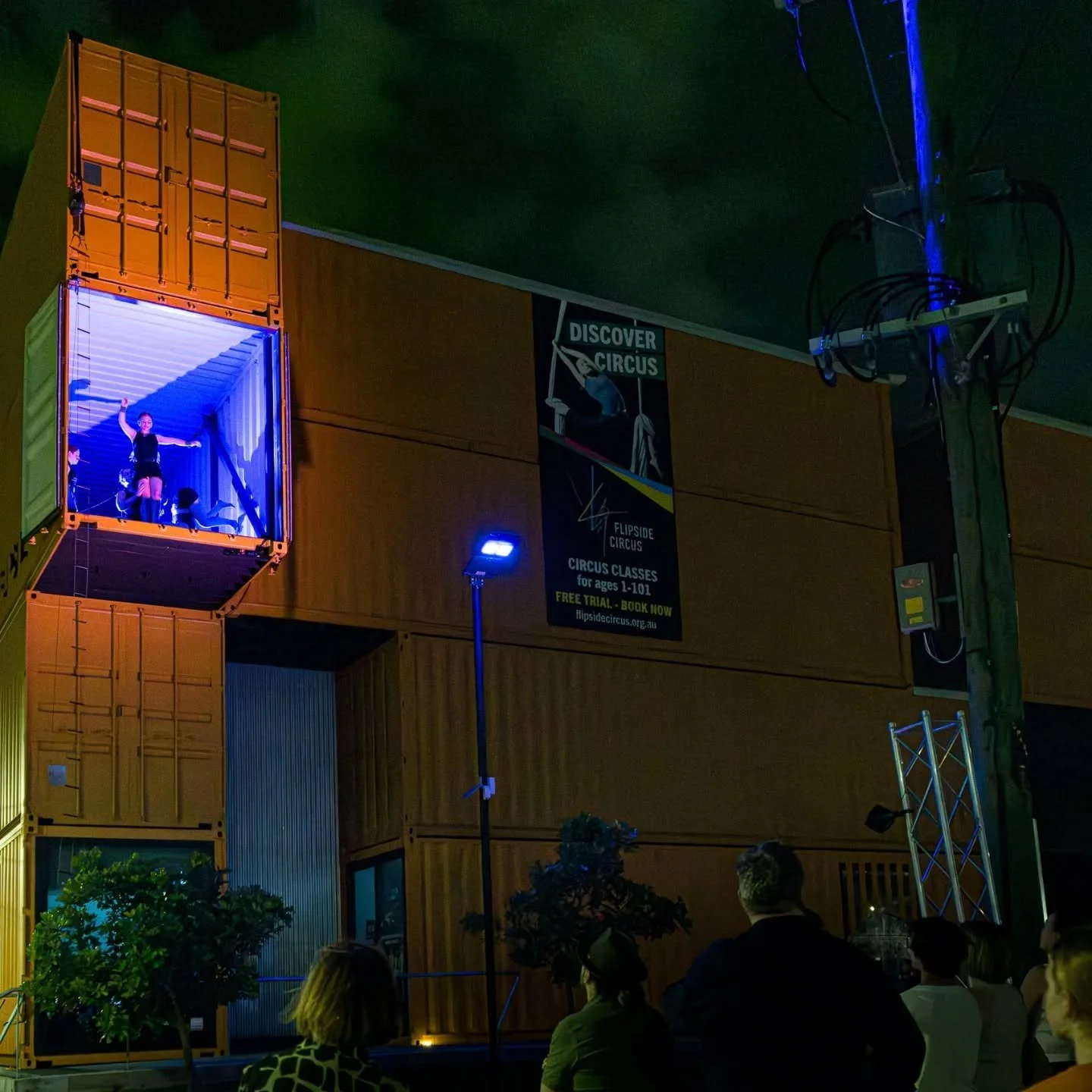 A circus performer inside a shipping container stage, waving to the audience at a circus event, with audience members seated outside in a dark venue.