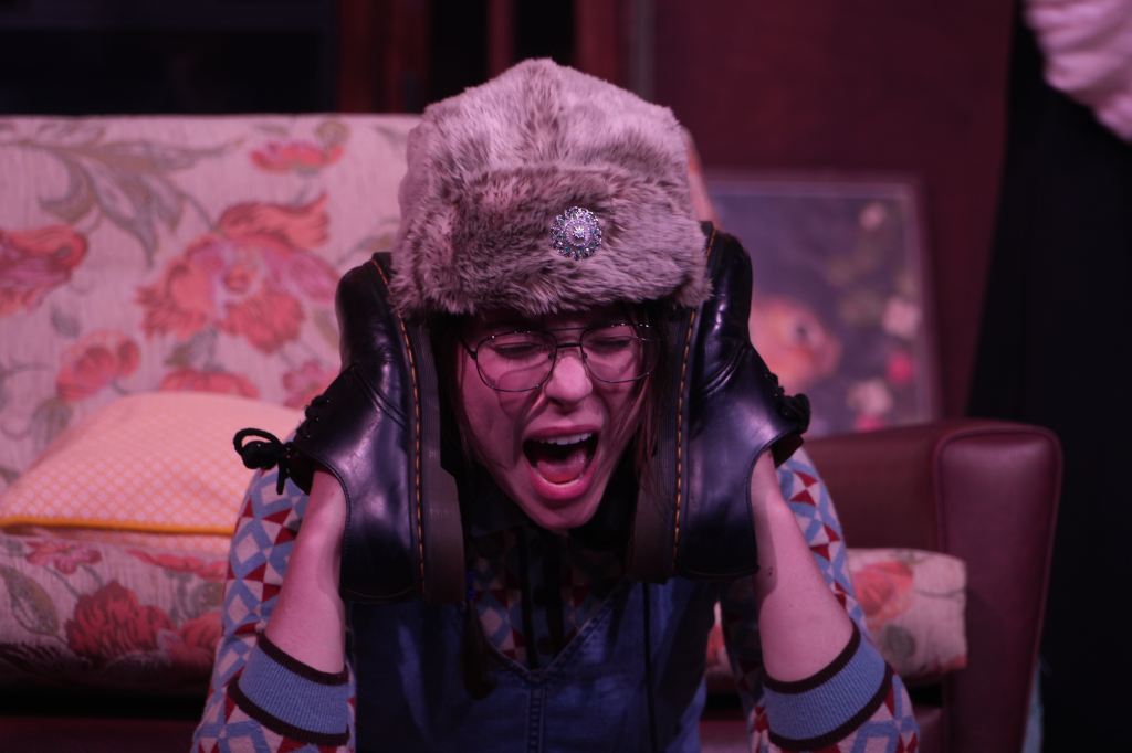 Woman wearing a fluffy hat with a brooch, glasses, and leather gloves, covering her ears and crying in a room with floral and patterned cushions.