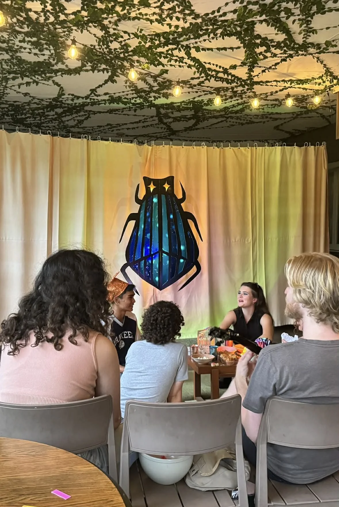 People gathered around a woman sitting at a table, with a colorful backdrop featuring a stylized insect with stars for eyes, in a decorated indoor space with string lights and greenery on the ceiling.