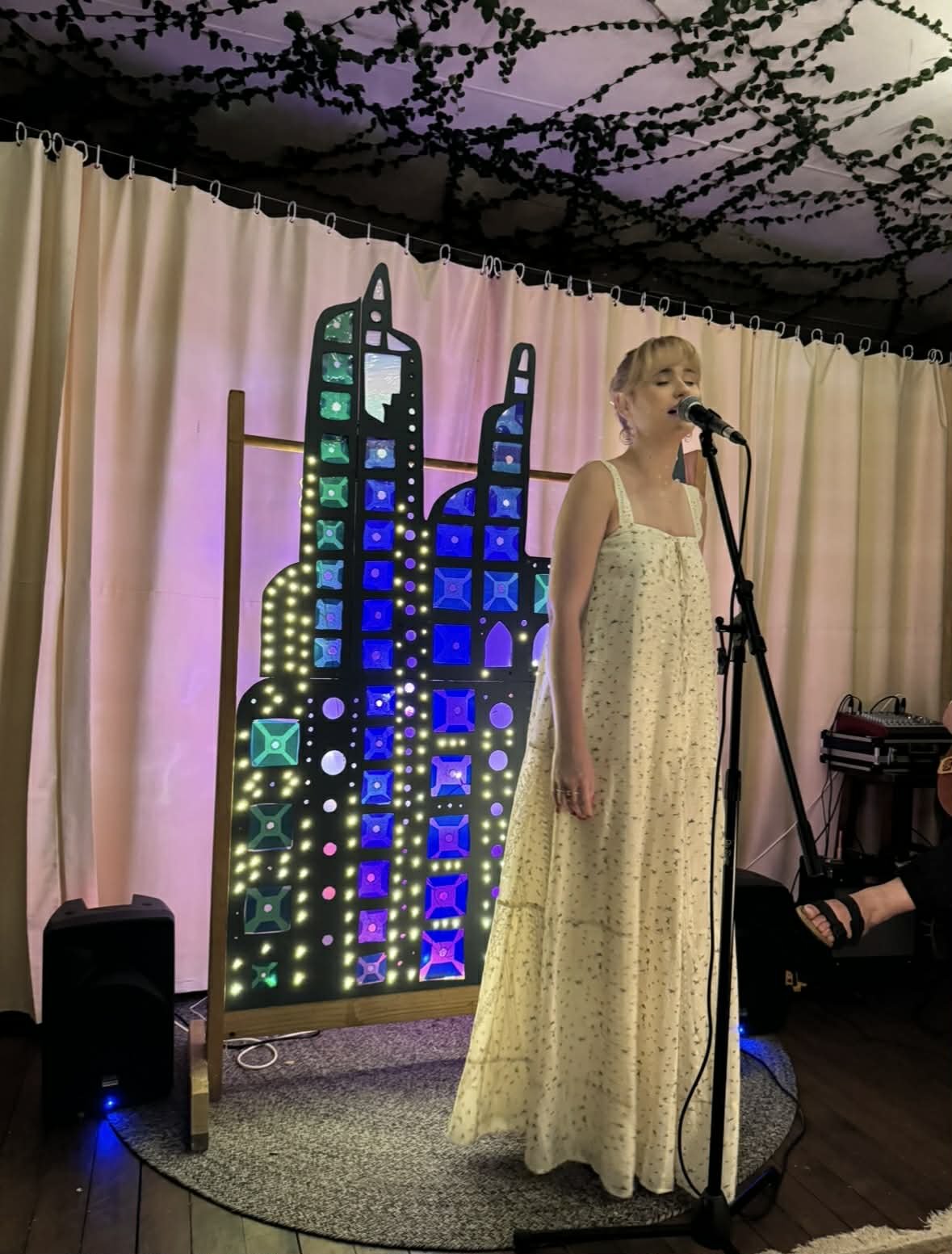 A woman singing into a microphone on stage with a decorative backdrop featuring colorful lights and geometric patterns, and curtains in the background.