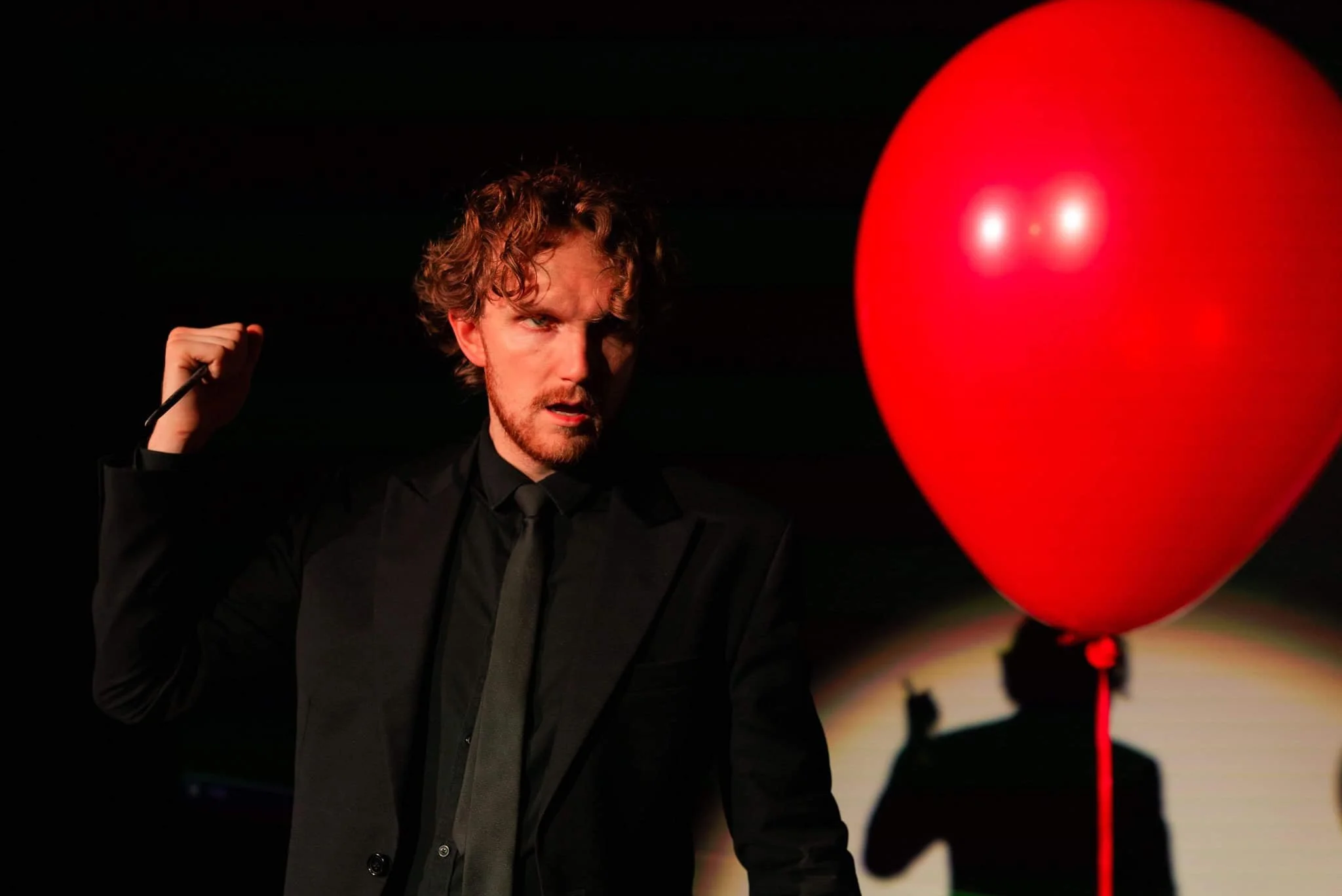 A man with curly hair and a beard dressed in black, holding a knife, looking intensely at a large red heart-shaped balloon.