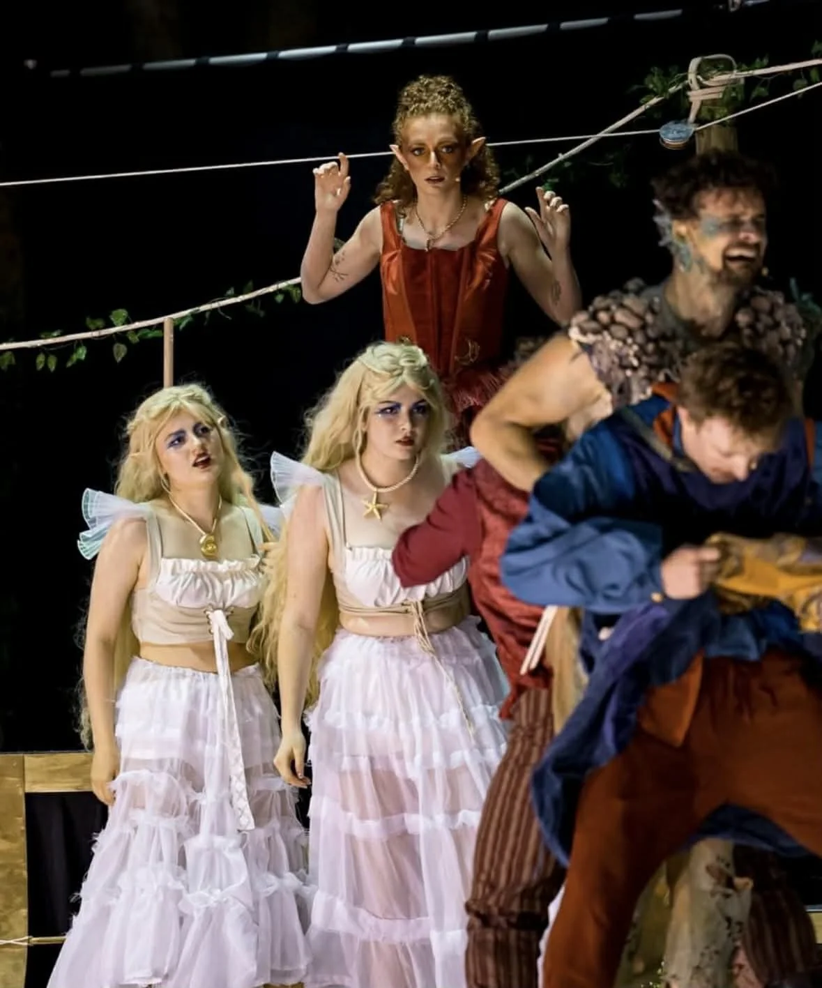 Scene from a theatrical play with actors in fantasy costumes, including fairy-like characters with blonde hair and fairy wings, and a woman with pointy ears and medieval attire, against a dark background with a rope and foliage, conveying a fantasy o