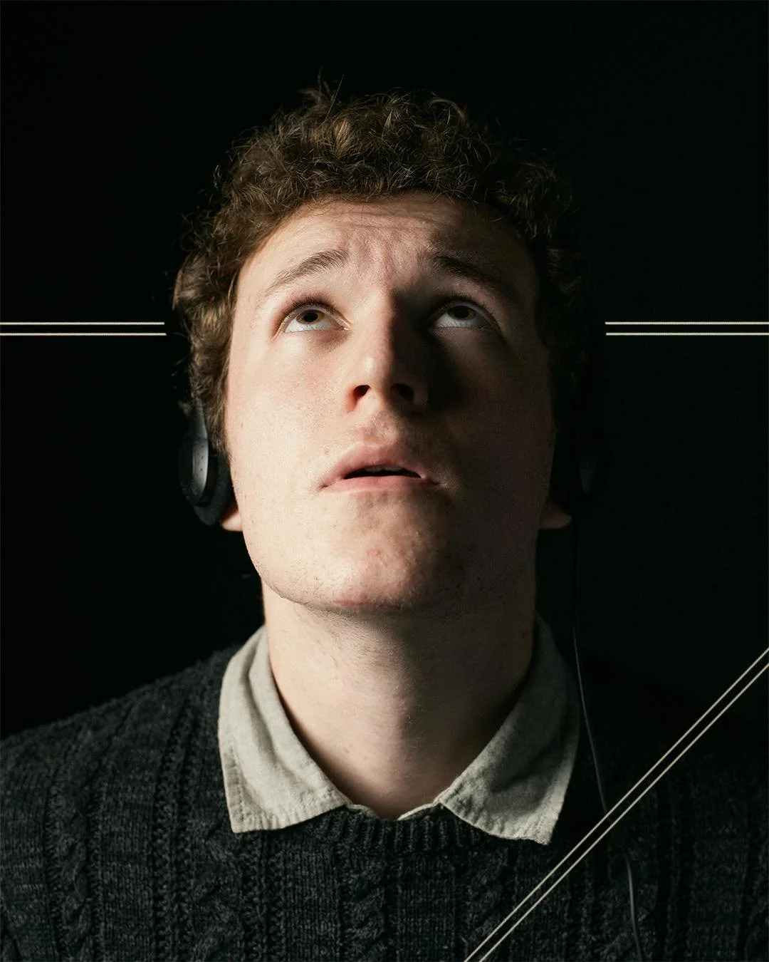 A young man with curly hair wearing headphones, looking upward against a black background with white lines.