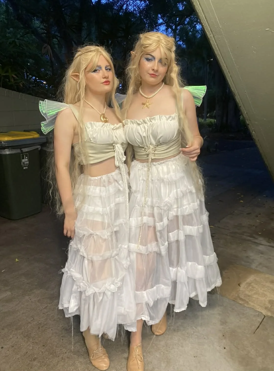 Two women dressed as mermaids standing outdoors at dusk, wearing cream-colored tops and long, flowy skirts with ruffled layers, elf ears, and glowing green wings on their shoulders. They have long blond hair, blue makeup, and are posing for the photo