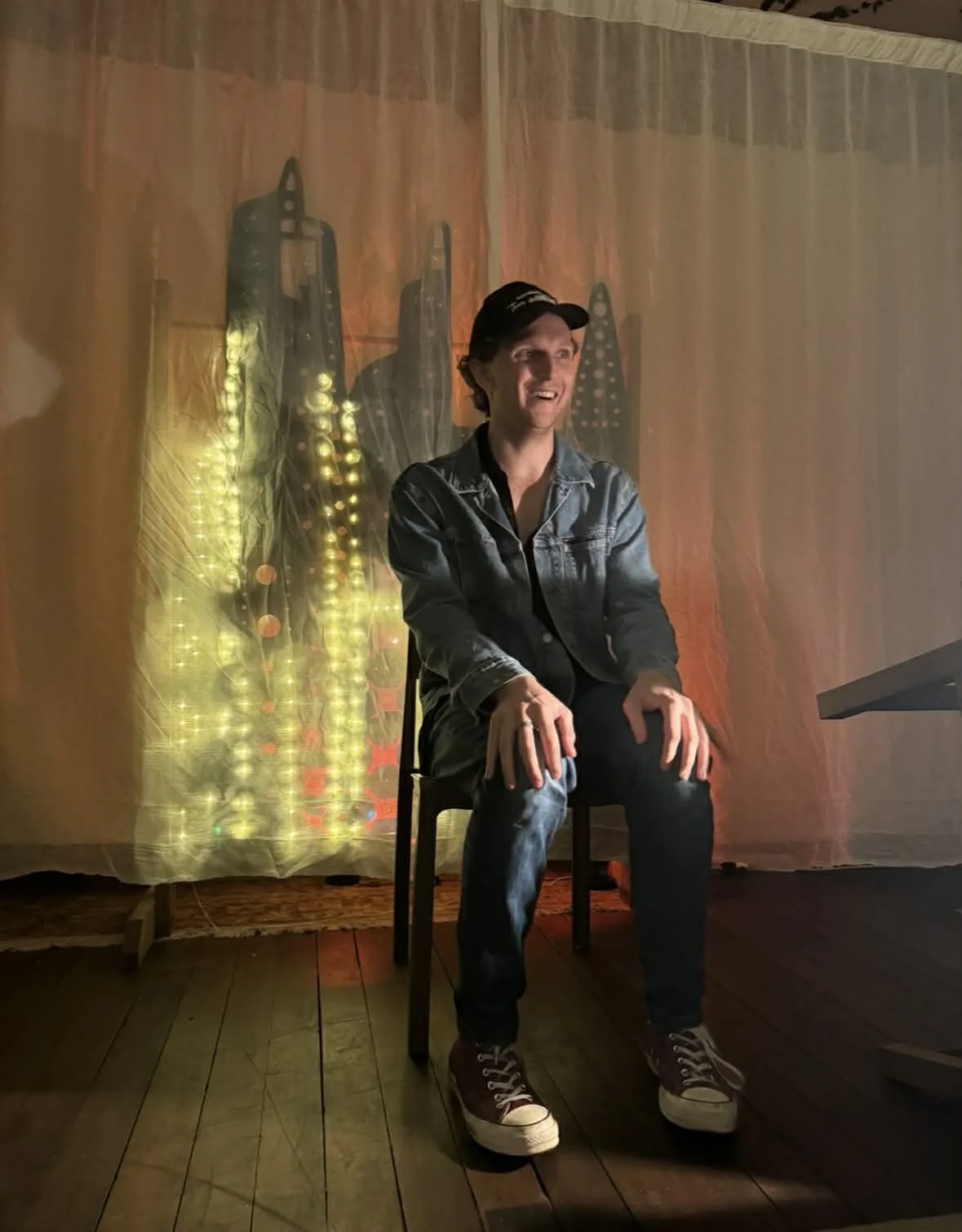 A man sitting on a chair in a dimly lit room with a curtain background and decorative hanging lights.