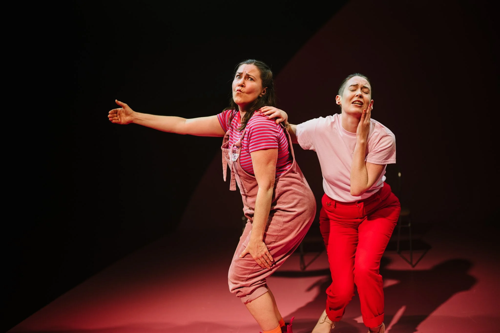 Two women on stage performing a comedic sketch or skit. One woman has her arm extended outward, and the other woman is touching her face with a concerned expression. Both are dressed casually in pink and red tones.