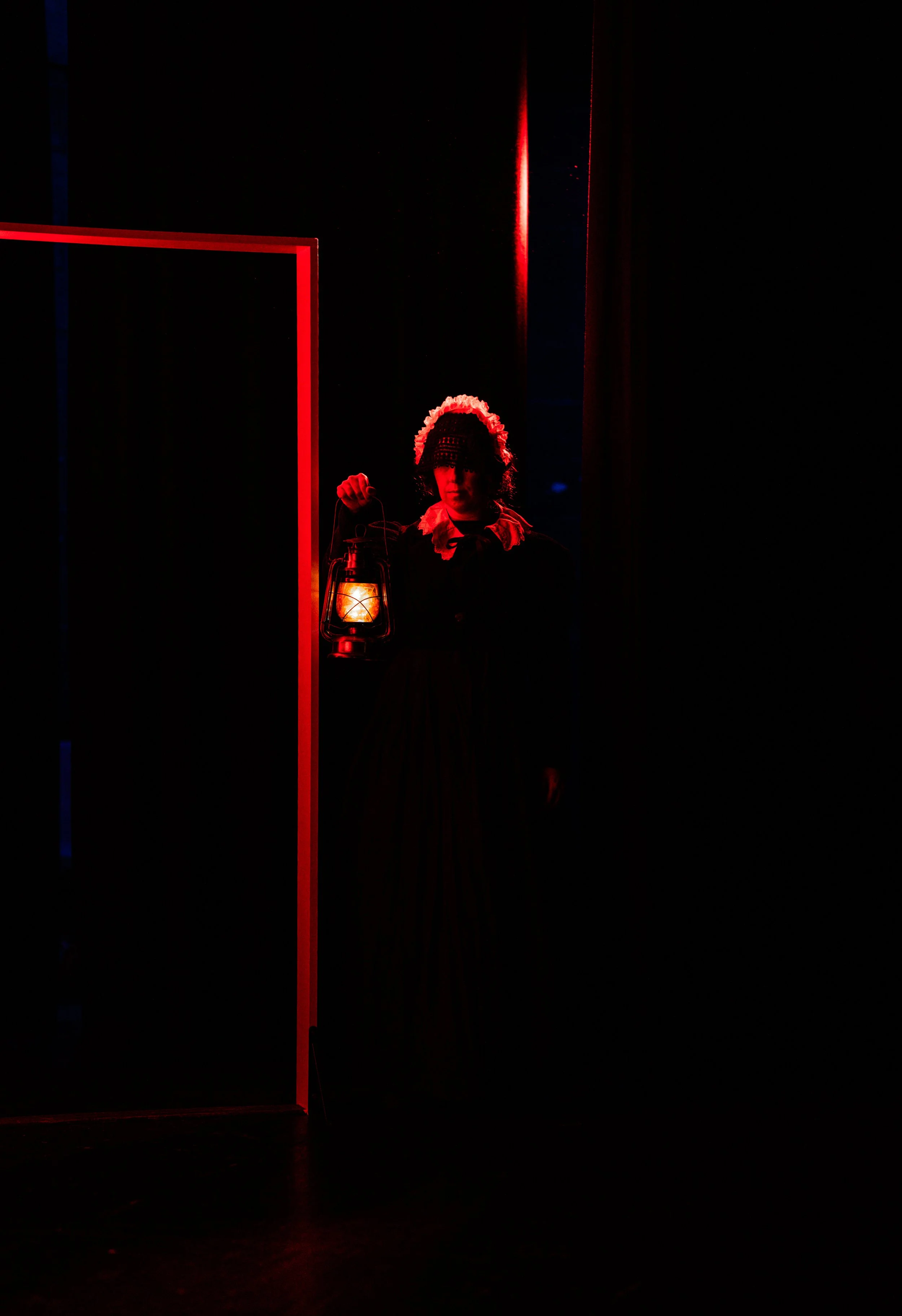 A woman dressed in vintage clothing holding a lantern, standing in a dark room with red lighting.