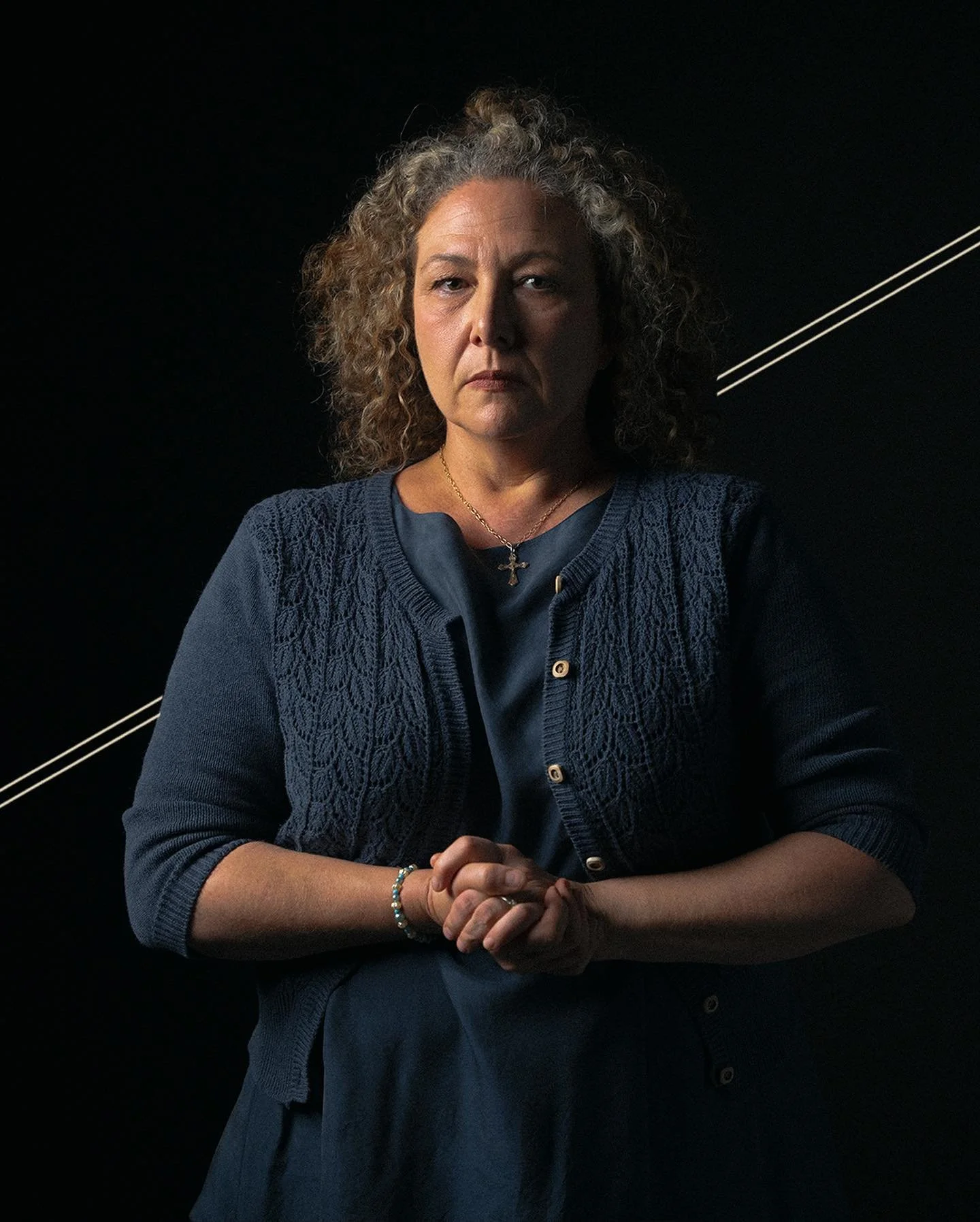 A middle-aged woman with curly gray hair stands against a dark background, wearing a navy blue cardigan over a dress, with her hands clasped in front of her. She has a serious expression, wearing a cross necklace and a bracelet.