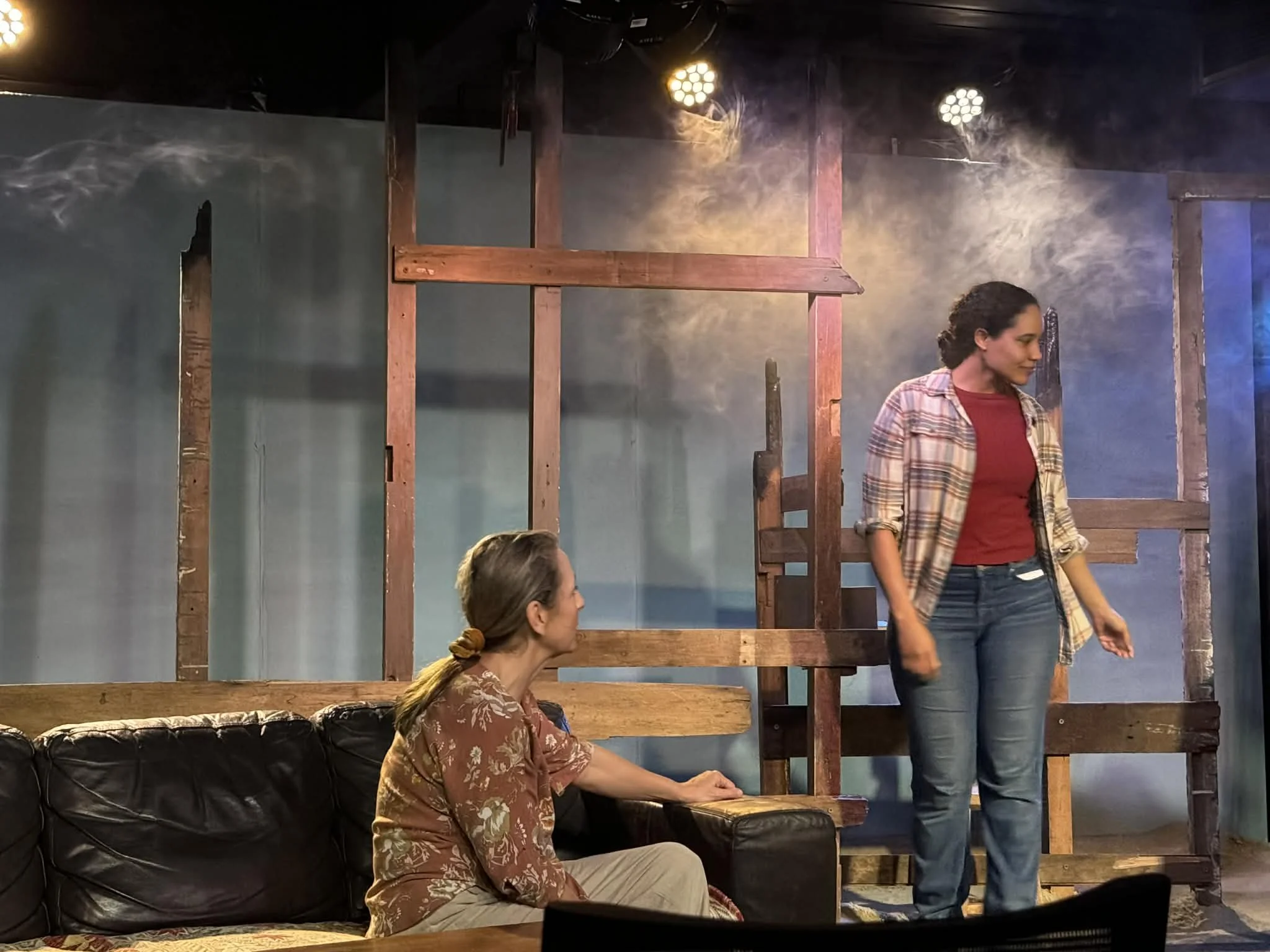 Two women in a room with a wooden framework and smoke, one sitting on a black sofa and the other standing nearby, engaged in conversation.