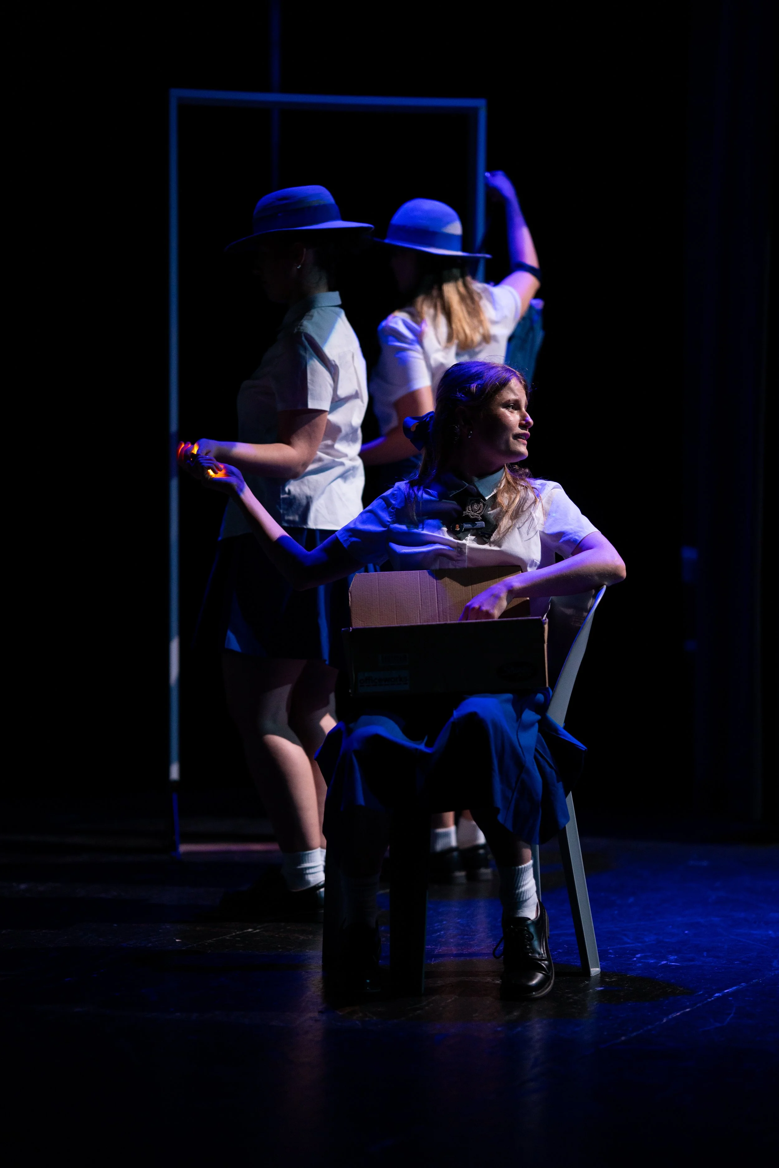 Three women on stage in school uniforms, with three standing and one seated, lit with dramatic blue lighting, performing in a theatrical production.