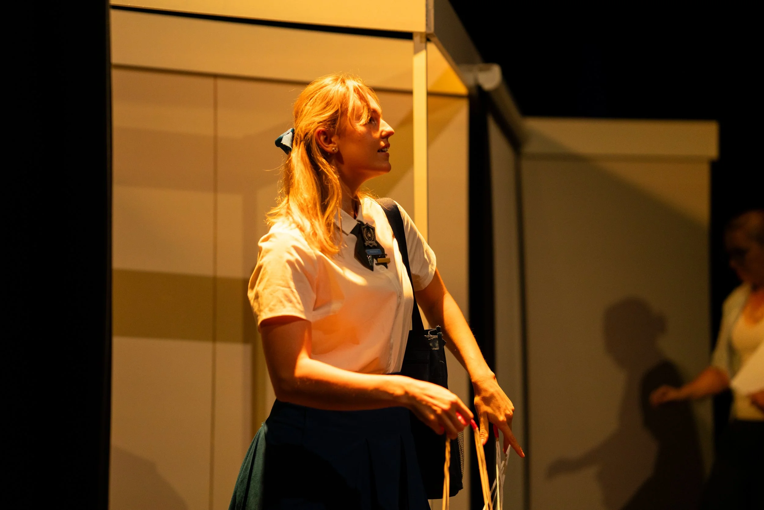A young woman with blonde hair tied back with a blue ribbon, wearing a cream-colored shirt and a dark skirt, standing on stage with a yellow and beige background. She is looking to her right, holding a long object and has a black bag over her shoulde