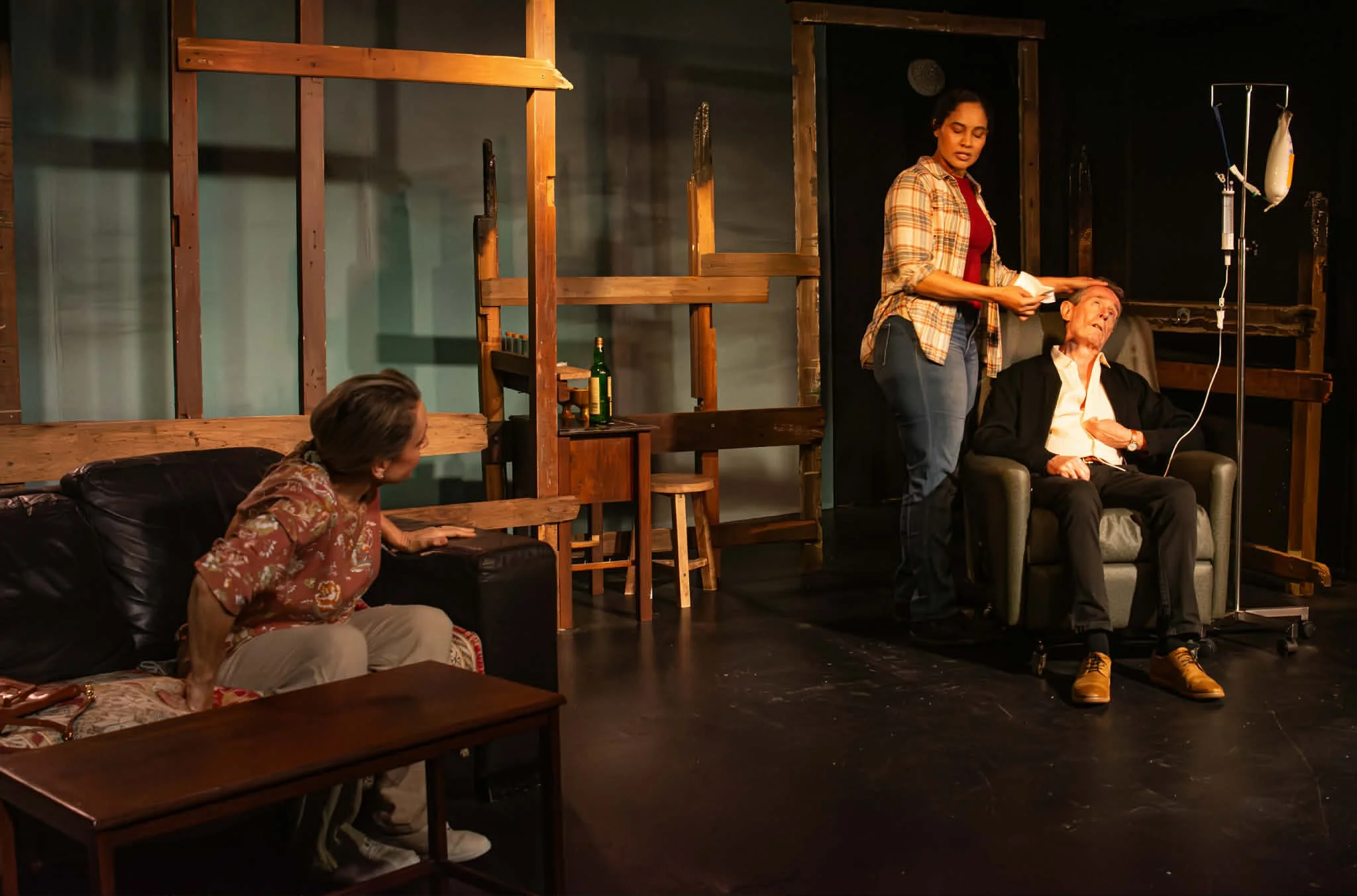 The scene depicts three women in a theatrical setting. One woman is seated on a black couch, leaning forward with her hand on the armrest, facing another woman who is seated in a chair and receiving medical care from a third woman standing beside her