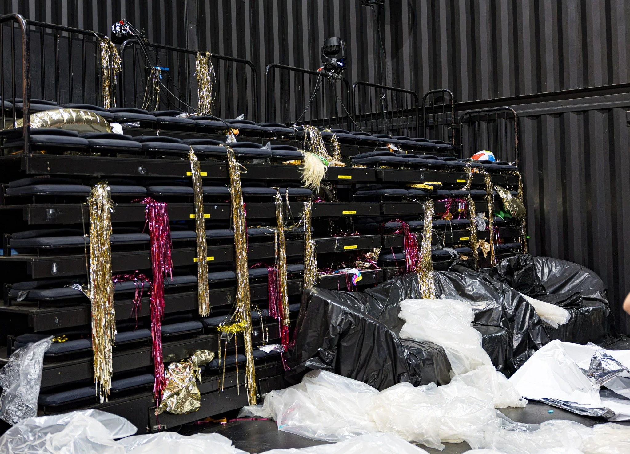 Empty black chairs decorated with gold and pink tinsel, with black, white, and silver wrapping paper and plastic sheets on the floor.