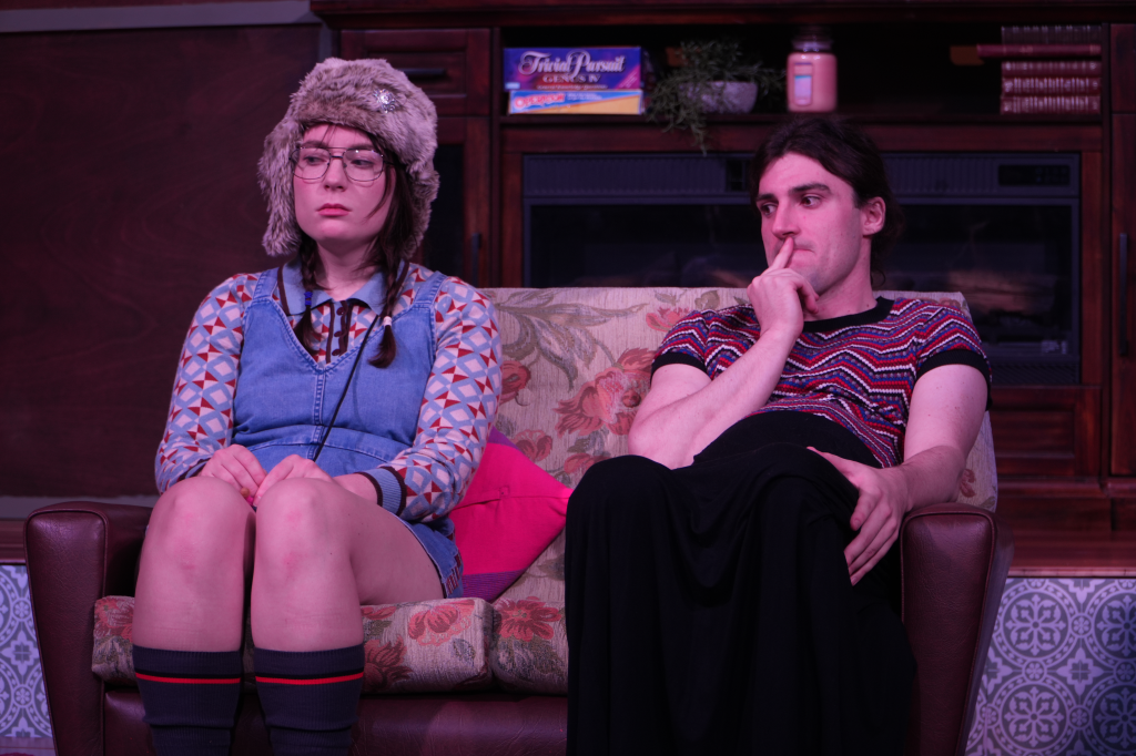 A young woman wearing a fuzzy hat, glasses, a patterned jacket, a denim pinafore, and knee-high socks sitting on a floral couch next to a young man in a colorful patterned shirt, who is sitting with his hand on his chin, appearing thoughtful. They ar