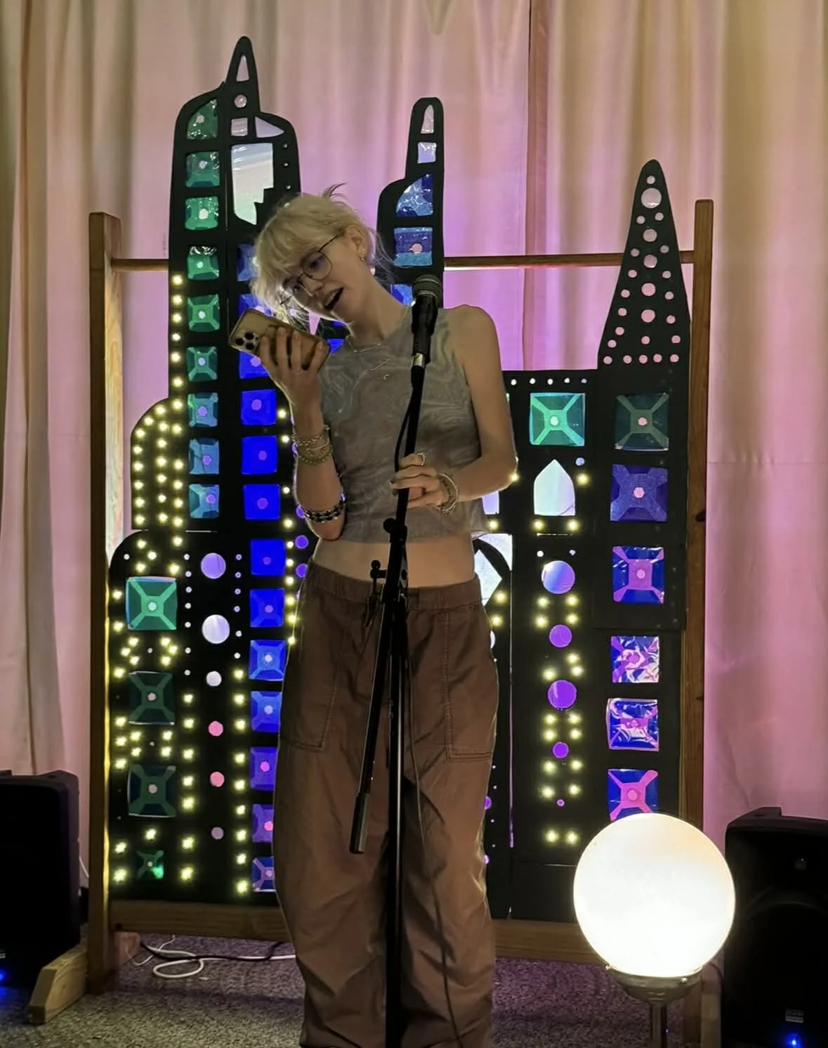 A woman with blonde hair and glasses is standing in front of a colorful LED cityscape backdrop. She is holding a phone and speaking into a microphone.