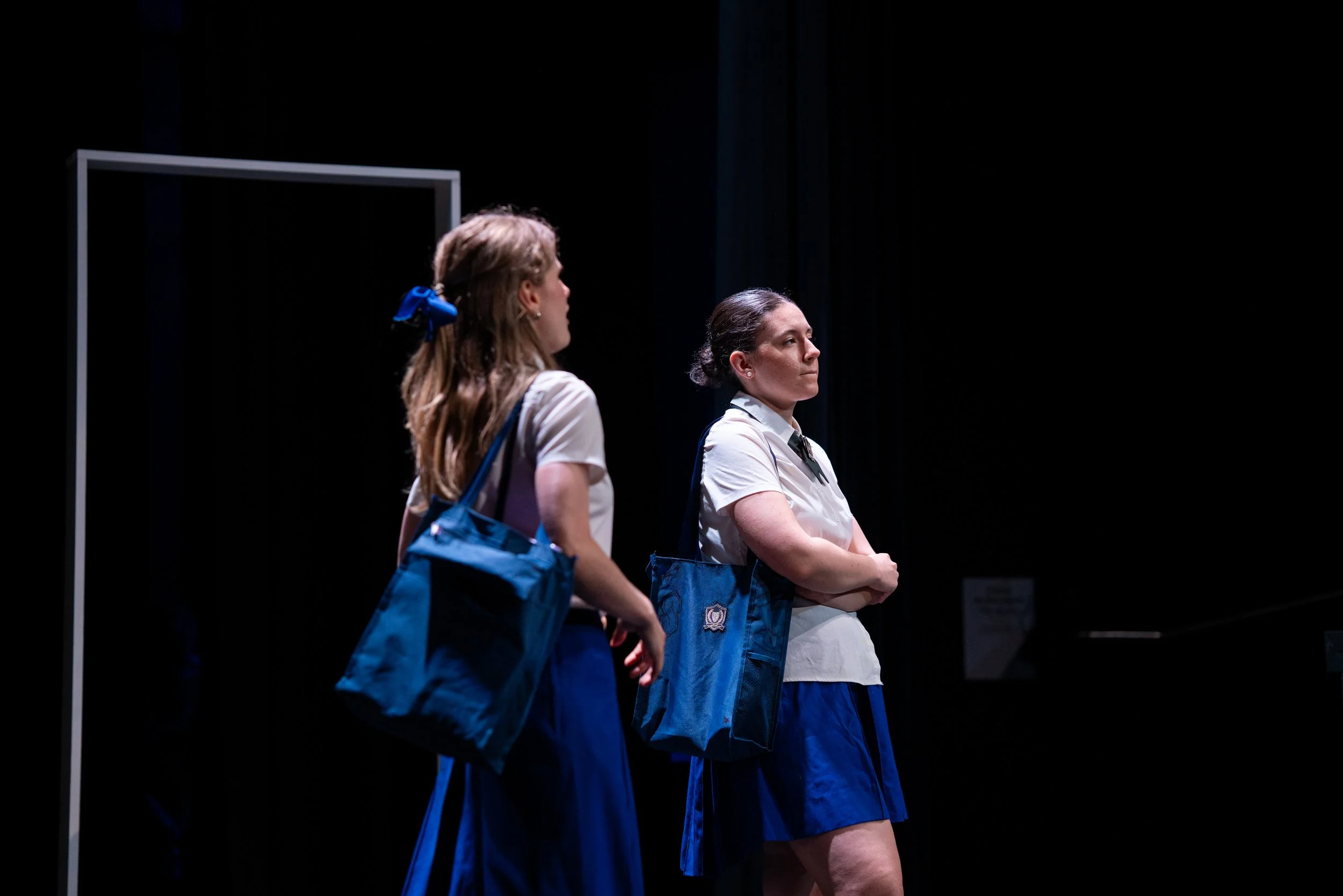 Two women in school uniforms standing on stage, facing forward with serious expressions, carrying blue bags.