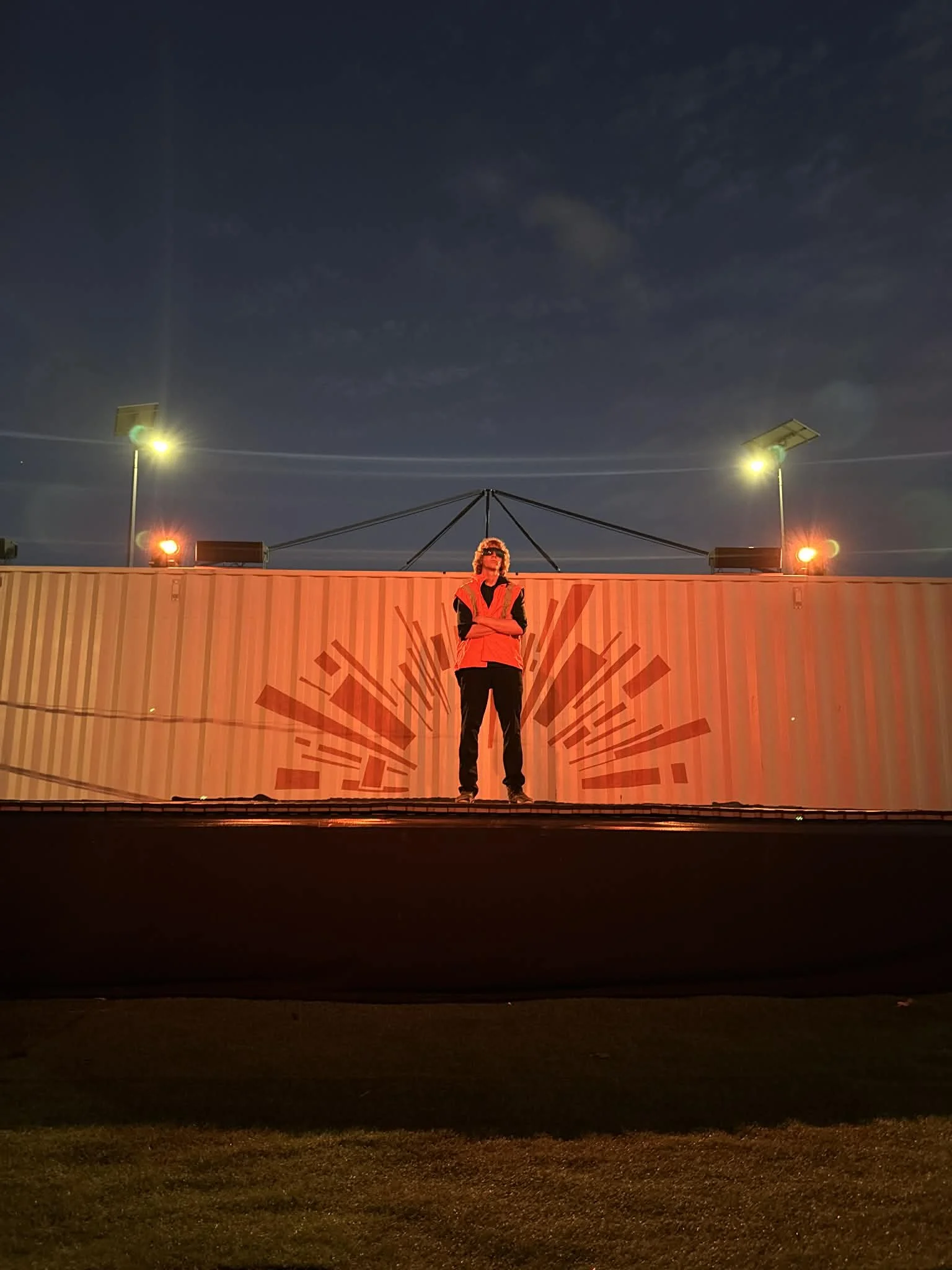 A person stands on a platform in front of a large shipping container with red geometric designs, illuminated by red and yellow lights at night.